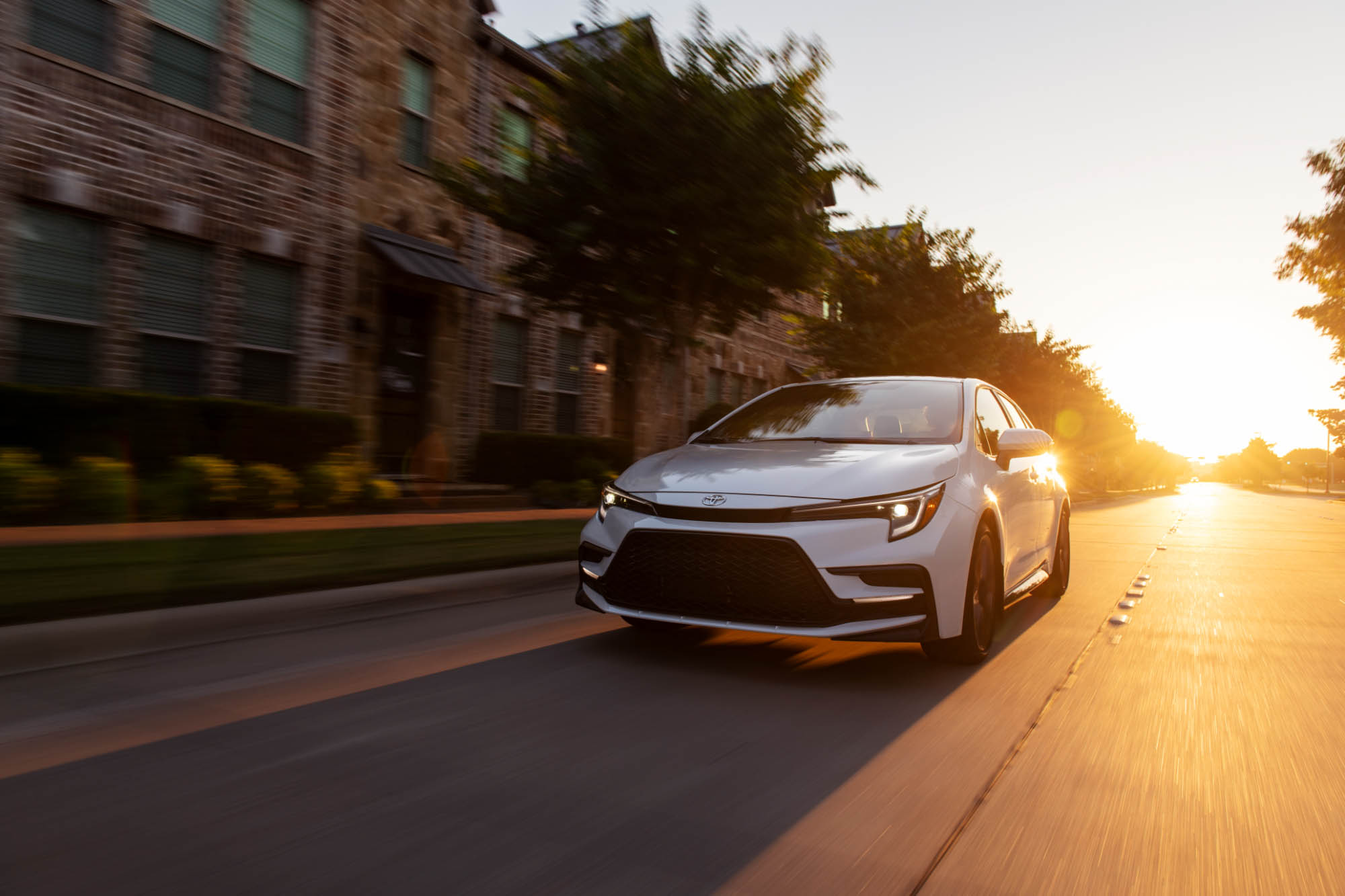 Silver Toyota Corolla driving in urban area with sun behind it.