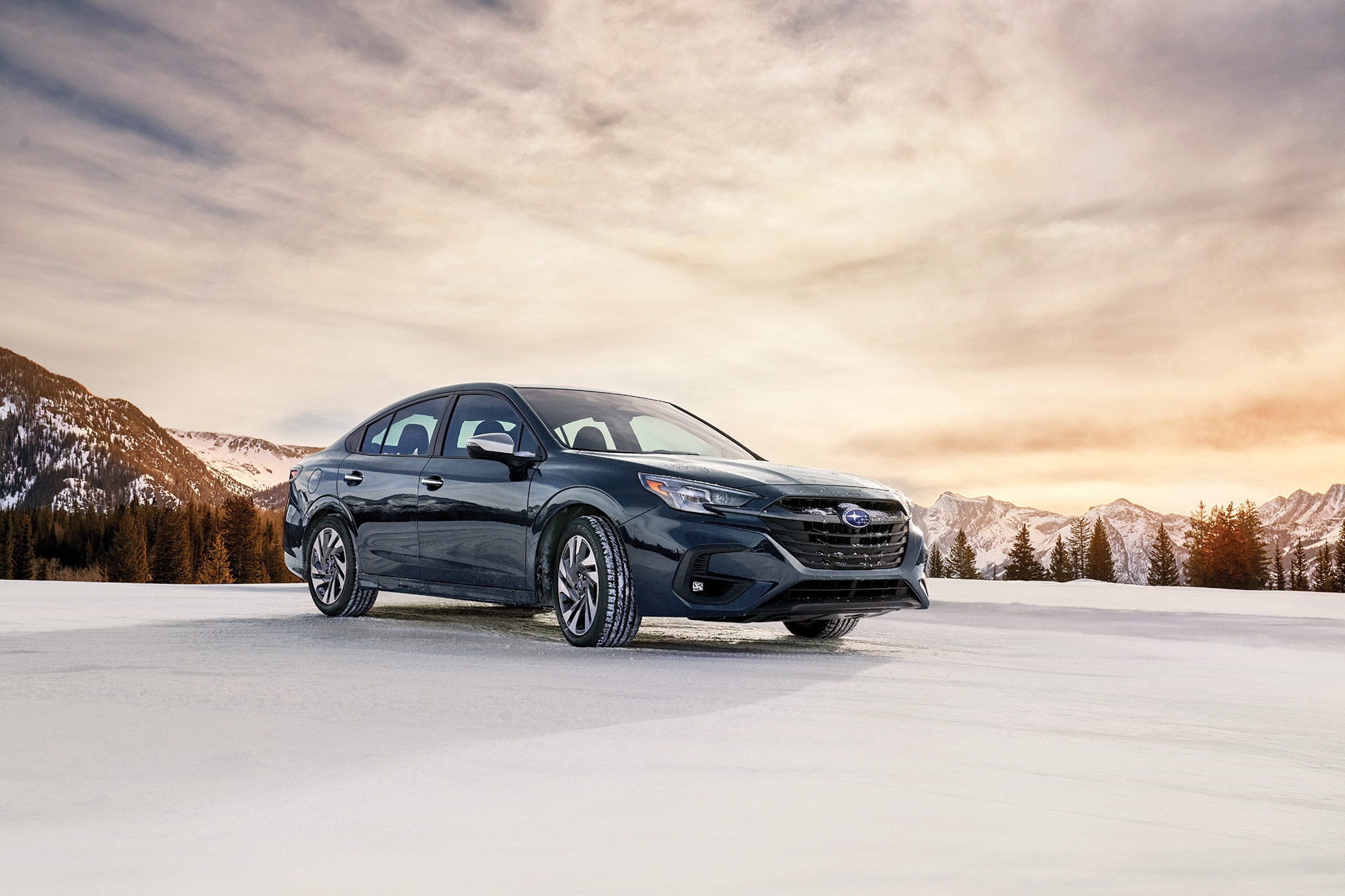 Black Subaru Legacy parked on snowy ground by mountains.