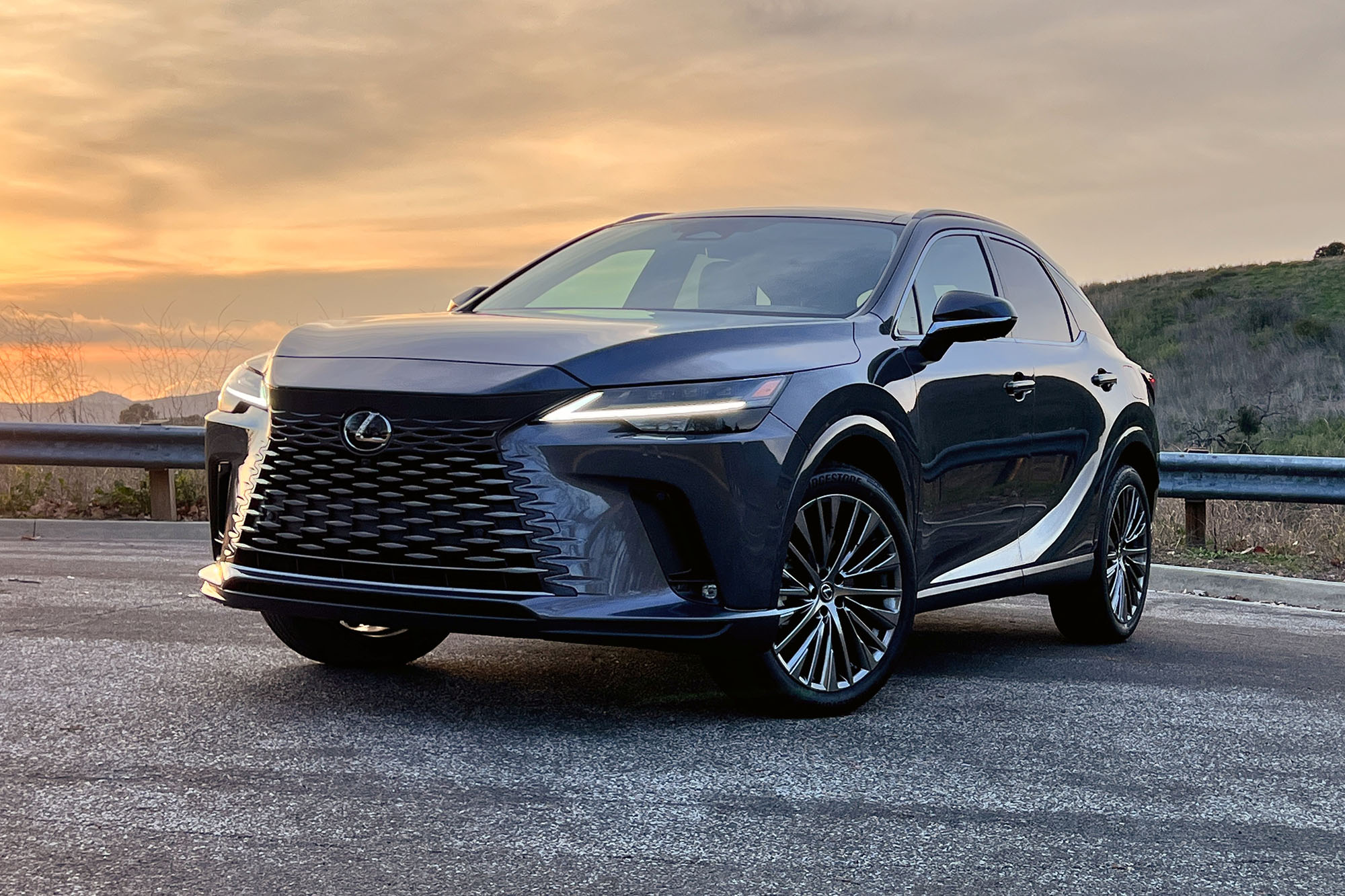 Front view of a dark gray 2024 Lexus RX 450h+ plug-in hybrid parked on the pavement at sunset.