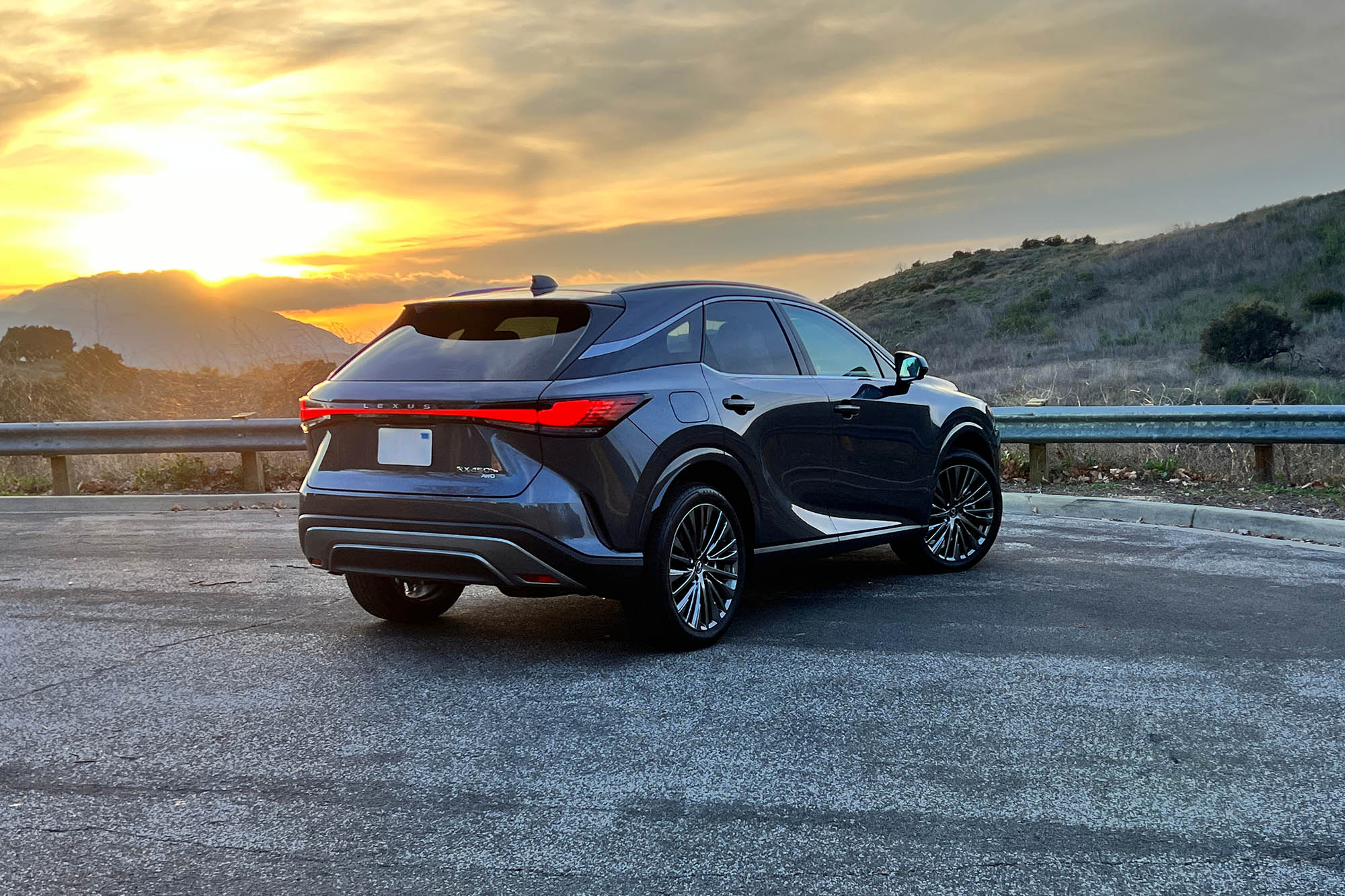 Rear view of a dark gray 2024 Lexus RX 450h+ plug-in hybrid parked on the pavement at sunset.