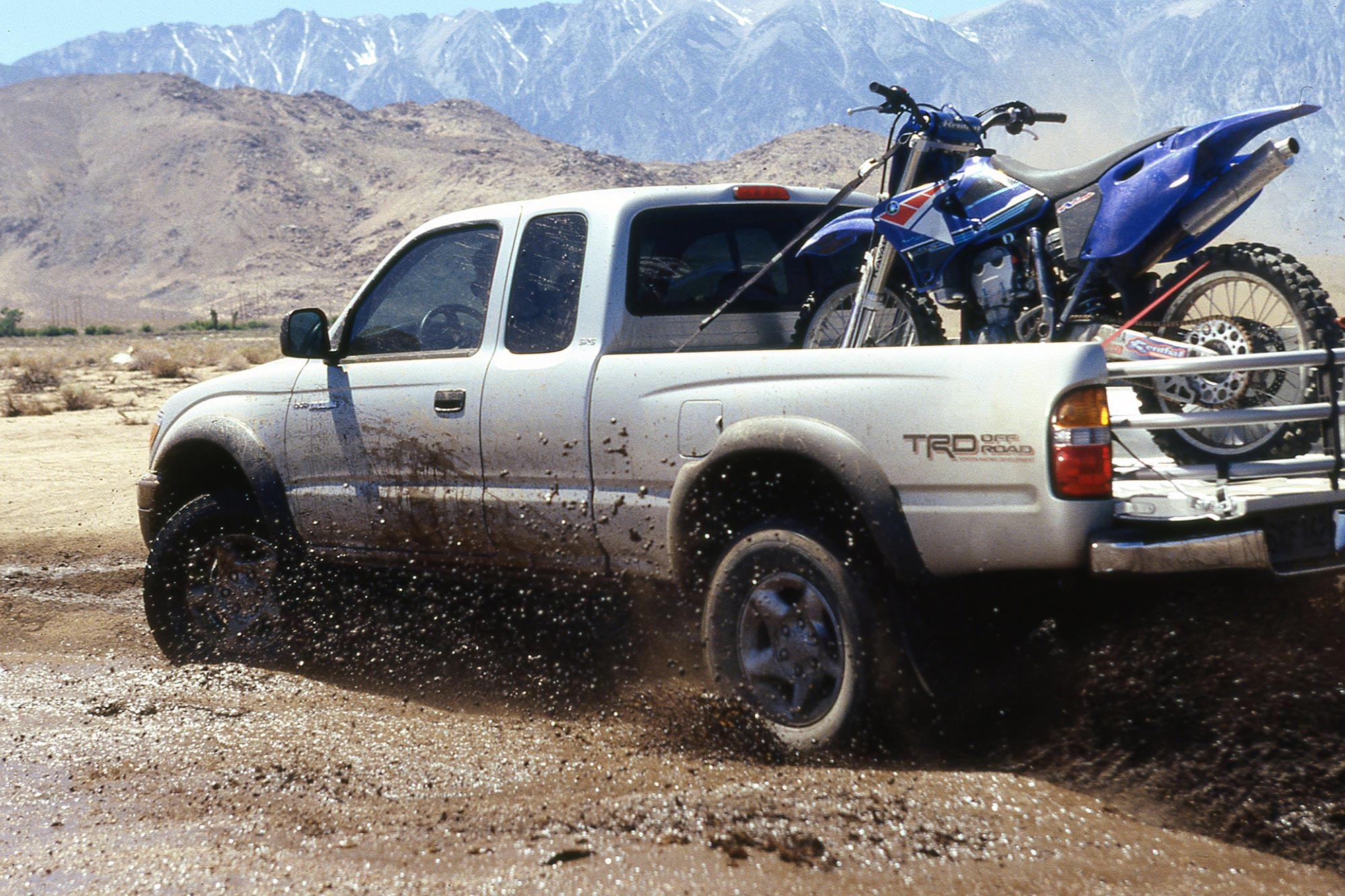 mid-1990s Toyota Tacoma driving through mud with motorcycle in bed.