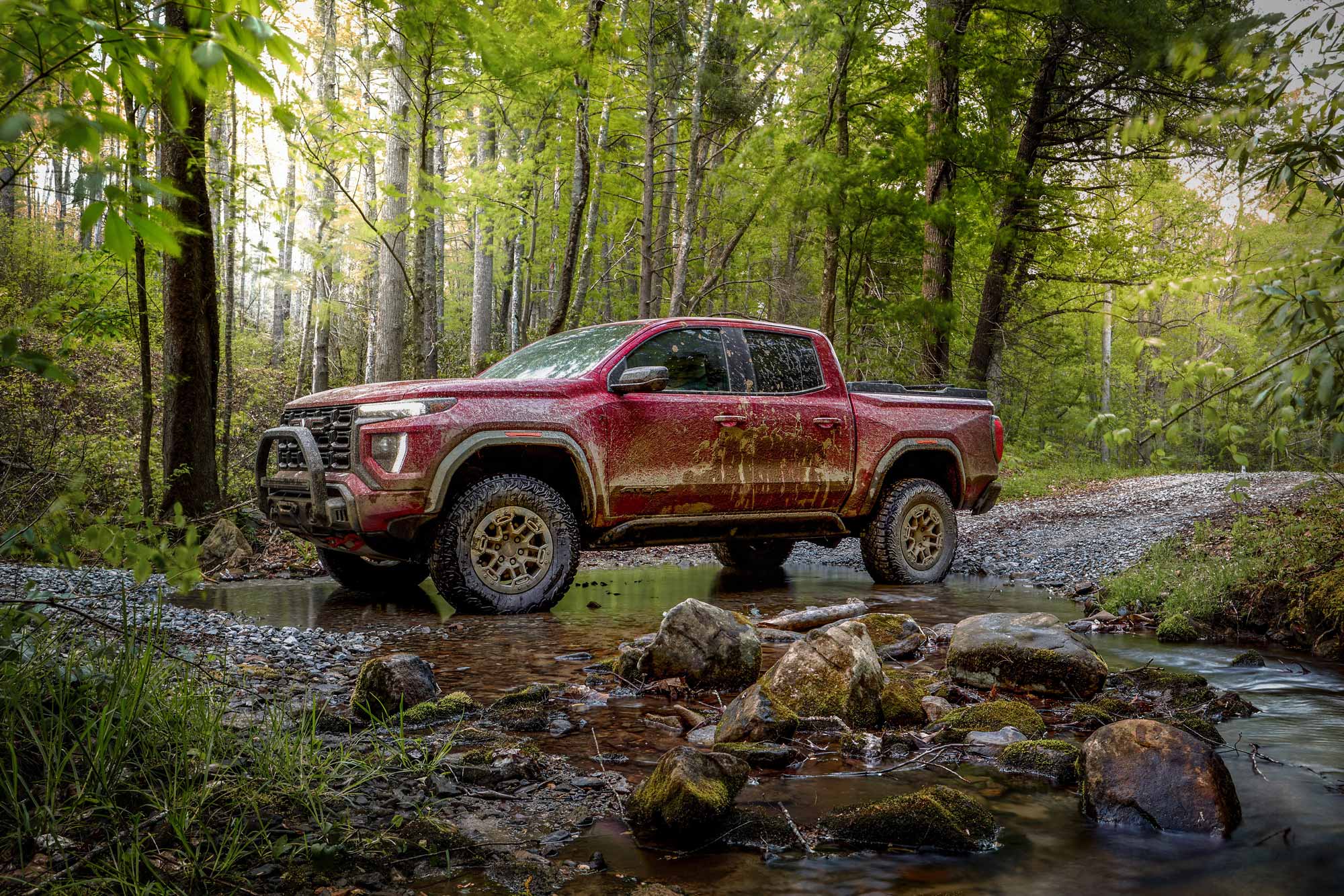 Red 2024 GMC Canyon AT4X covered in mud and parked in a shallow creek.
