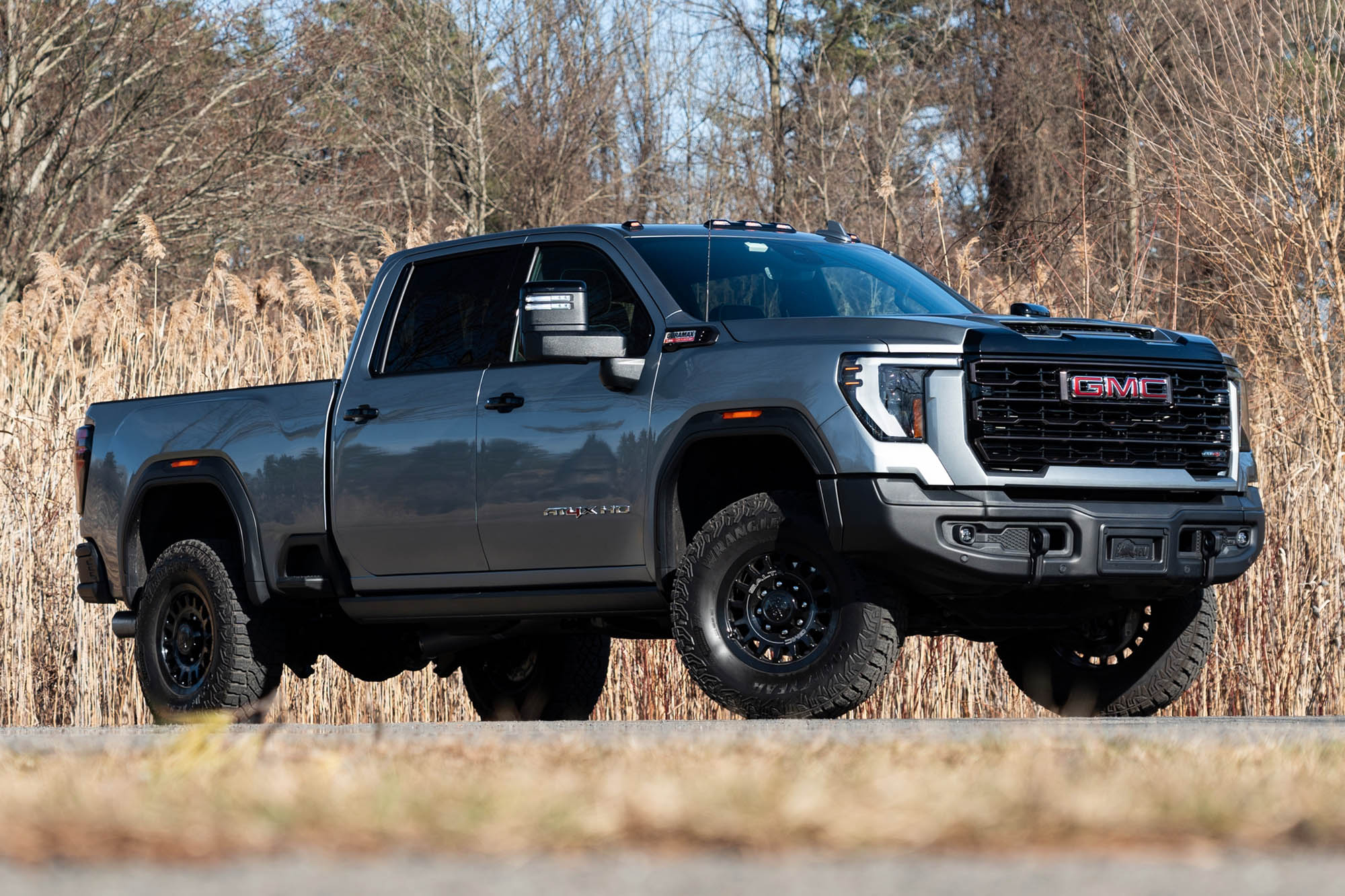 Front view of a gray 2024 GMC Sierra 2500HD with the AT4X AEV package parked on pavement near some trees.