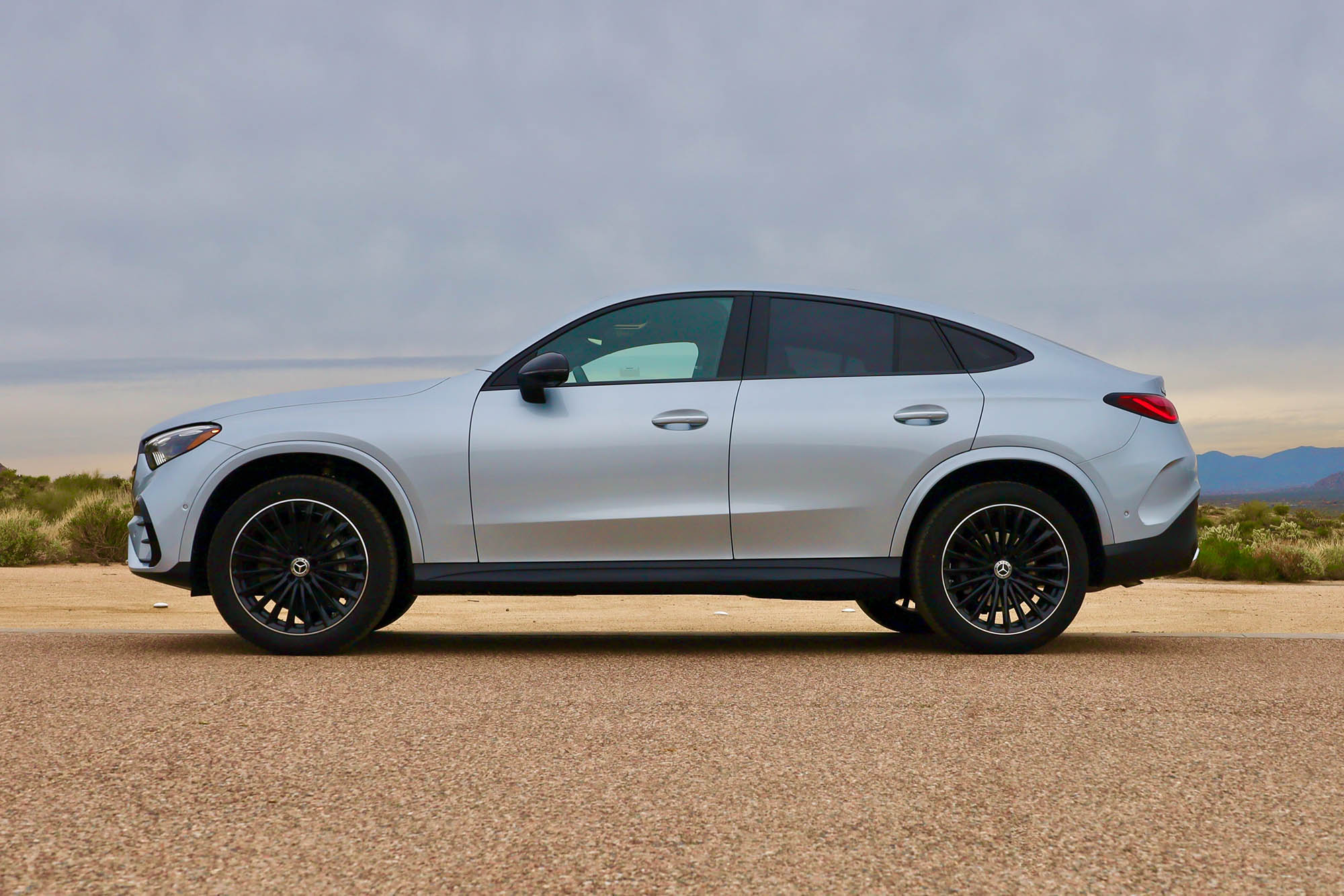 Side view of a silver 2024 Mercedes-Benz GLC 300 parked on the pavement in the Arizona desert with distant mountains and a gray sky in the background.