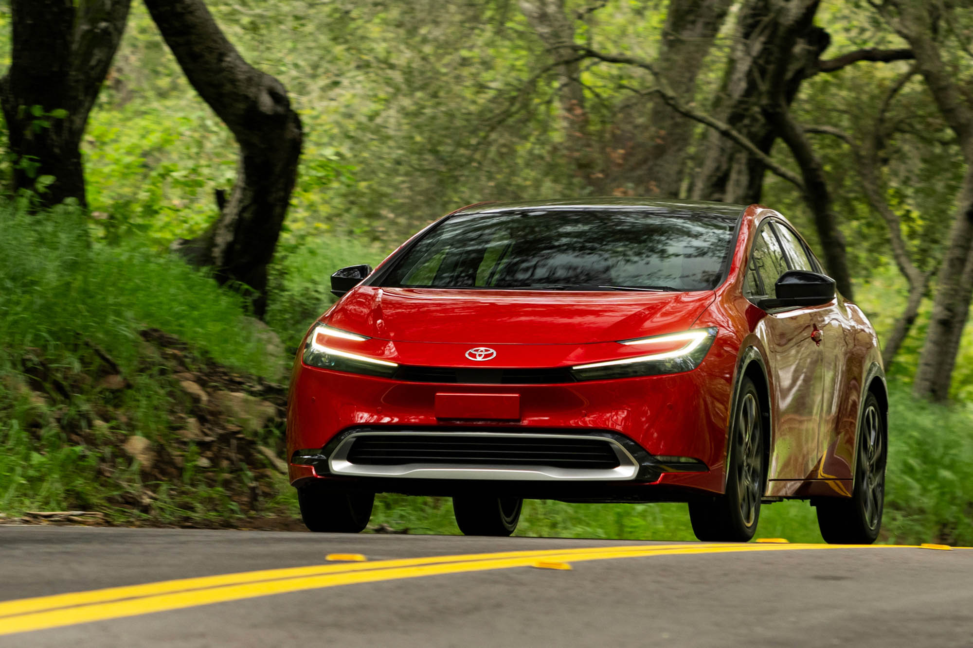Red 2024 Toyota Prius Prime driving on paved road with trees on the side.
