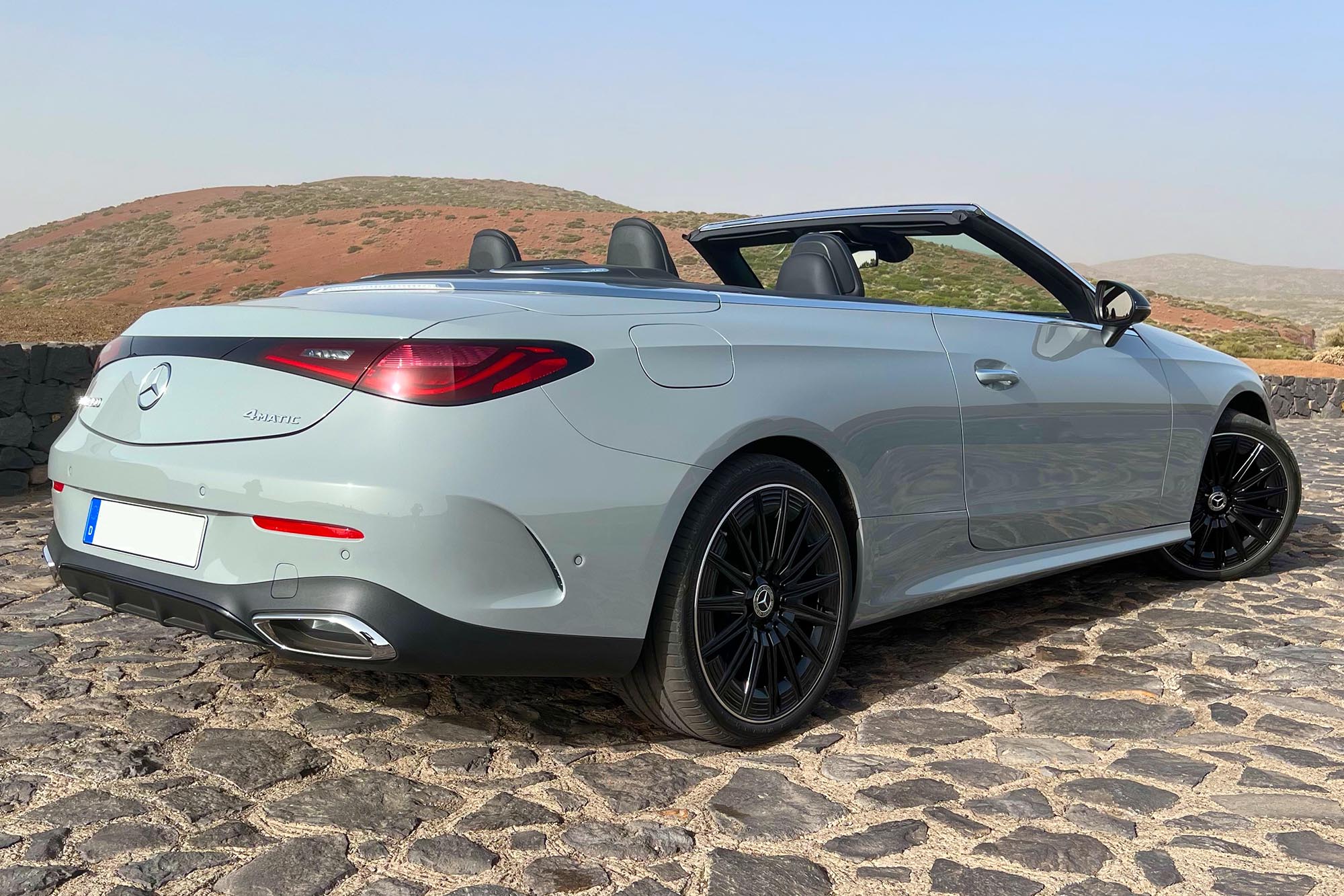 Rear-quarter view of a 2024 Mercedes-Benz CLE 450 Cabriolet, parked with the top down on a sunny day on a cobblestone surface.