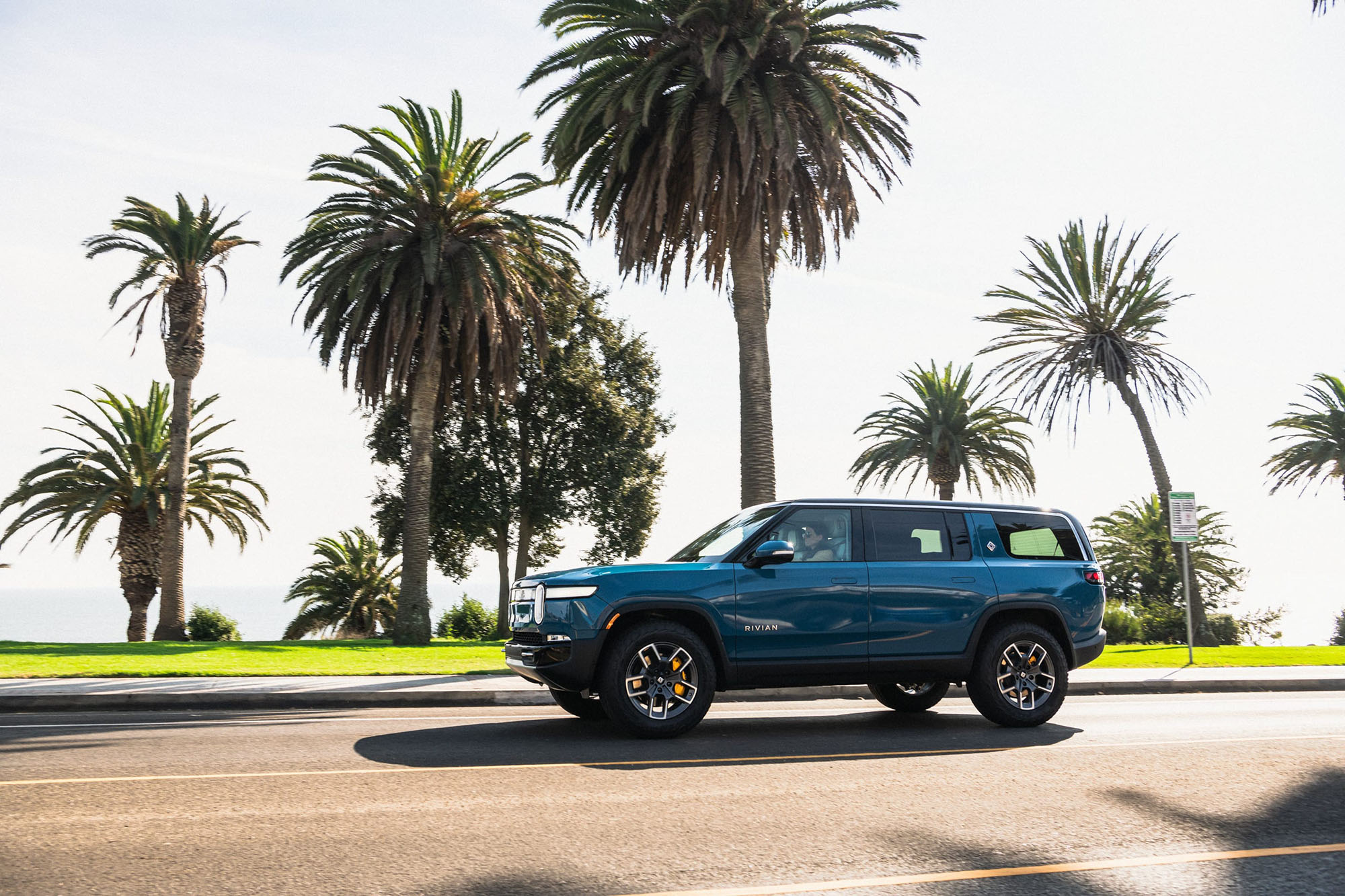 Rivian R1S in dark teal driving on road next to palm trees.