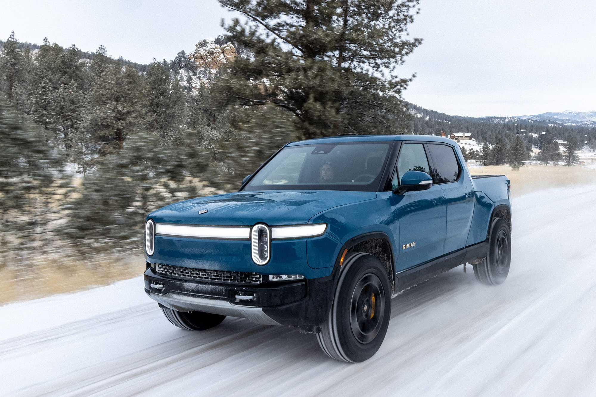 Rivian R1T in dark teal driving on snowy road.