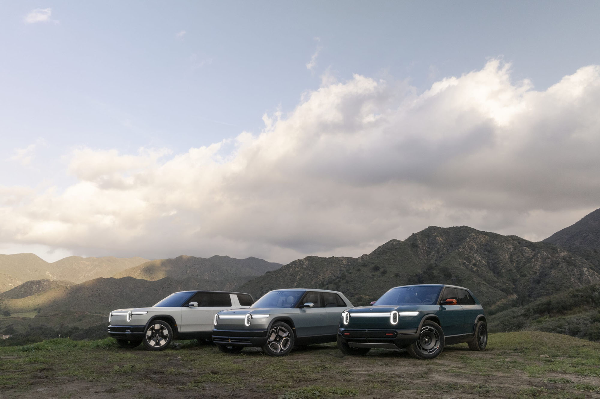 Rivian R1S, R1T, and R2 vehicles parked in front of mountains.