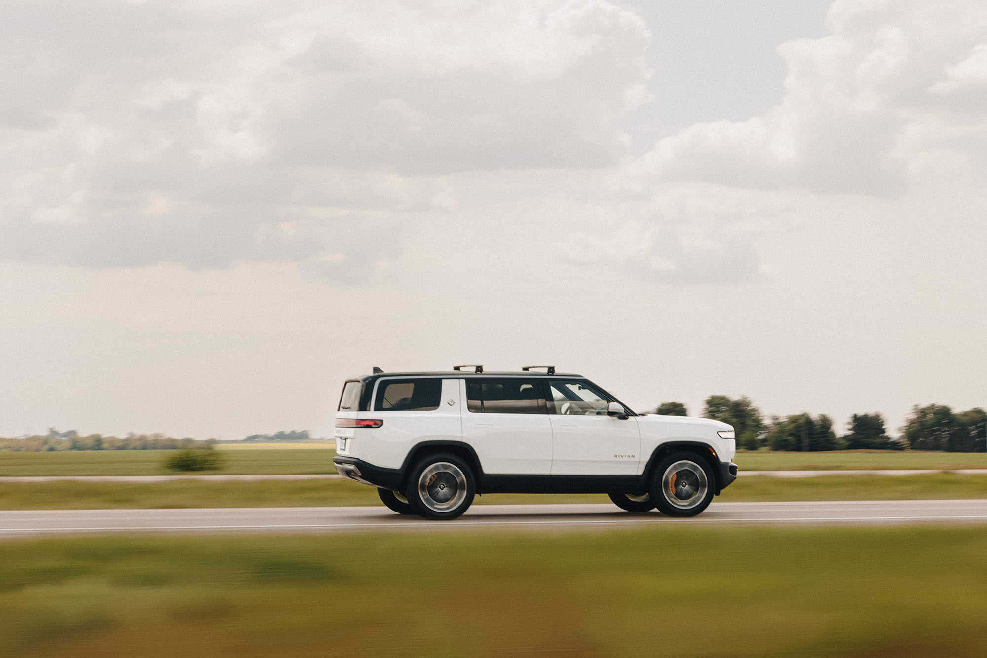 Rivian R1S in white driving on paved road with grass on either side.