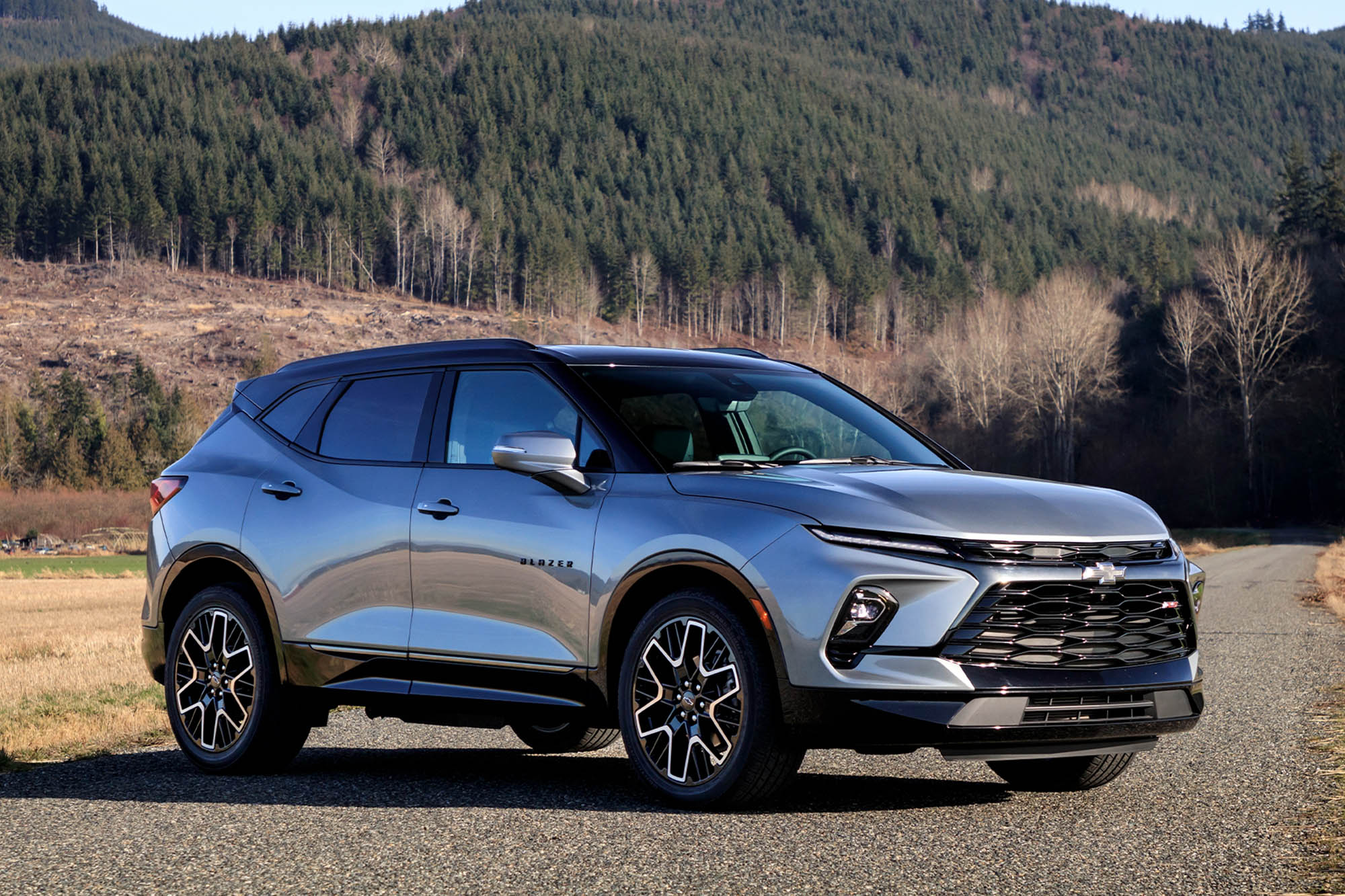 Text: Front quarter view of a silver 2024 Chevrolet Blazer RS parked on a road with a tree-covered mountain in the background.