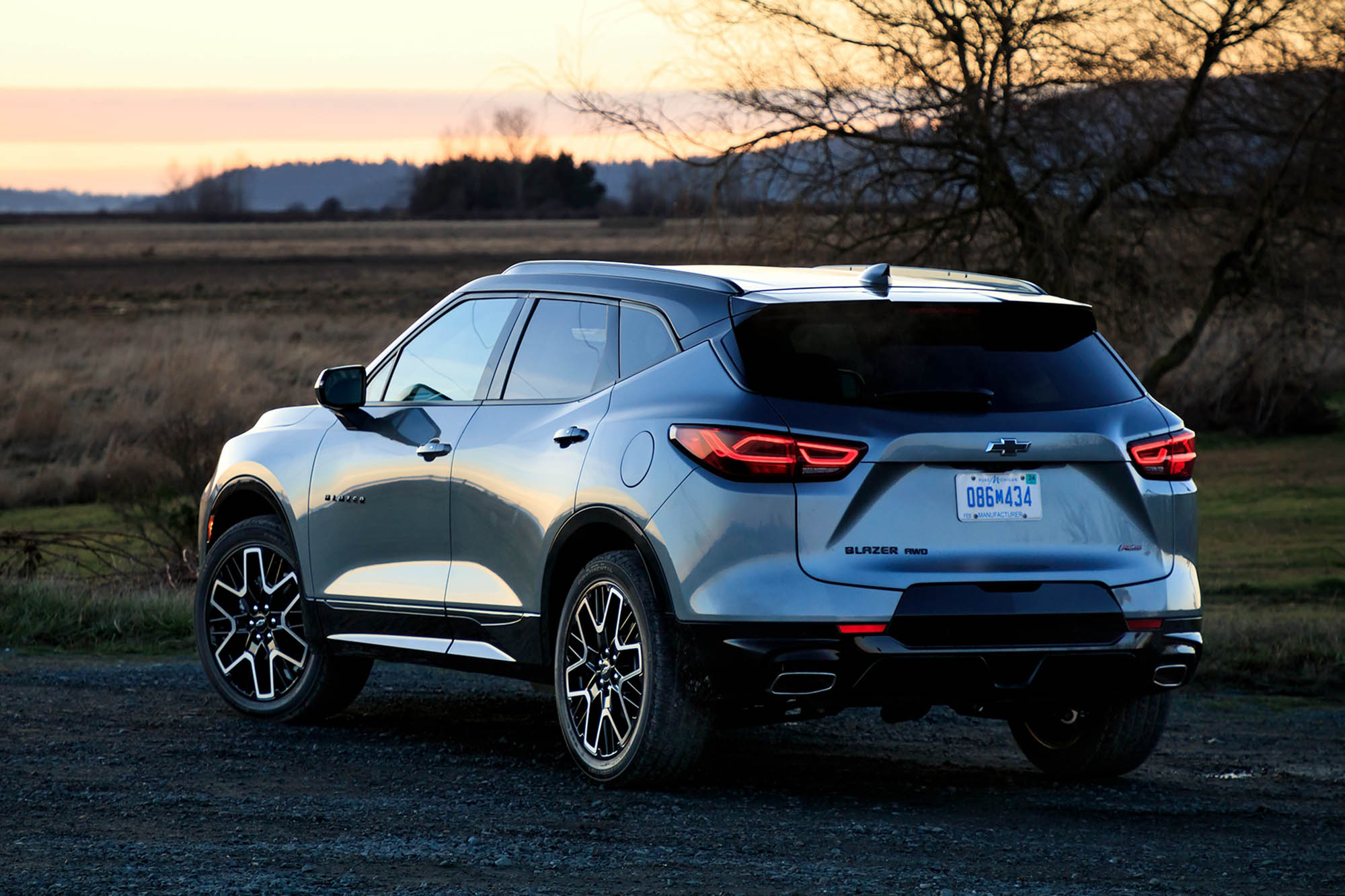 Rear quarter view of a silver 2024 Chevrolet Blazer RS parked on gravel at sunset with a field in the background.