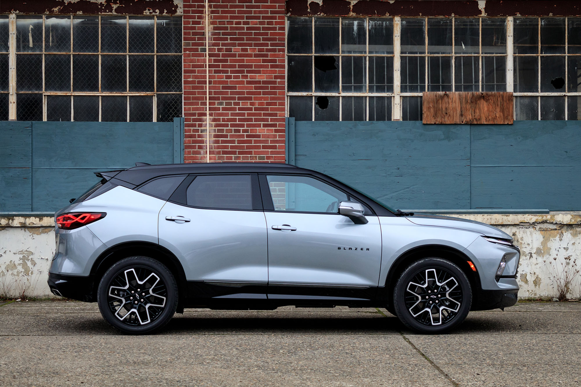 Side view of a silver 2024 Chevrolet Blazer RS parked on the pavement with an old industrial building in the background.