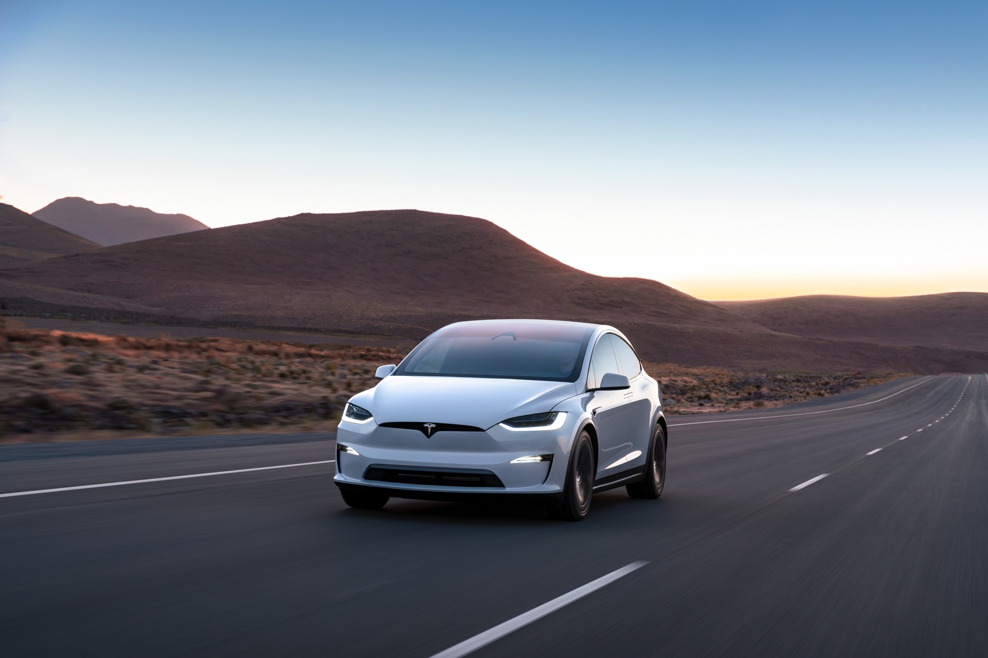 White Tesla Model X driving down highway next to hilltops