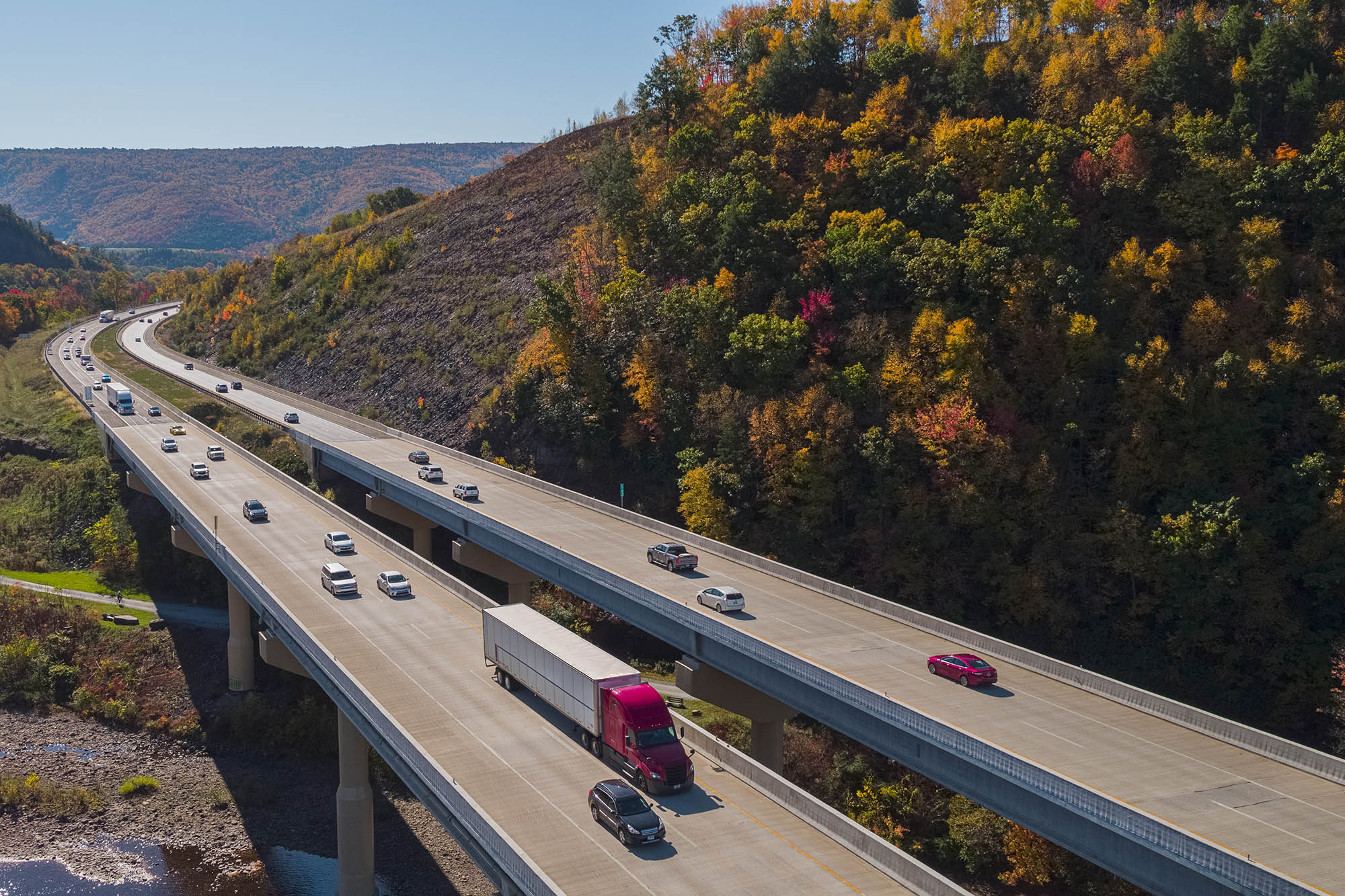 Cars and trucks drive on highway beside trees and mountains.