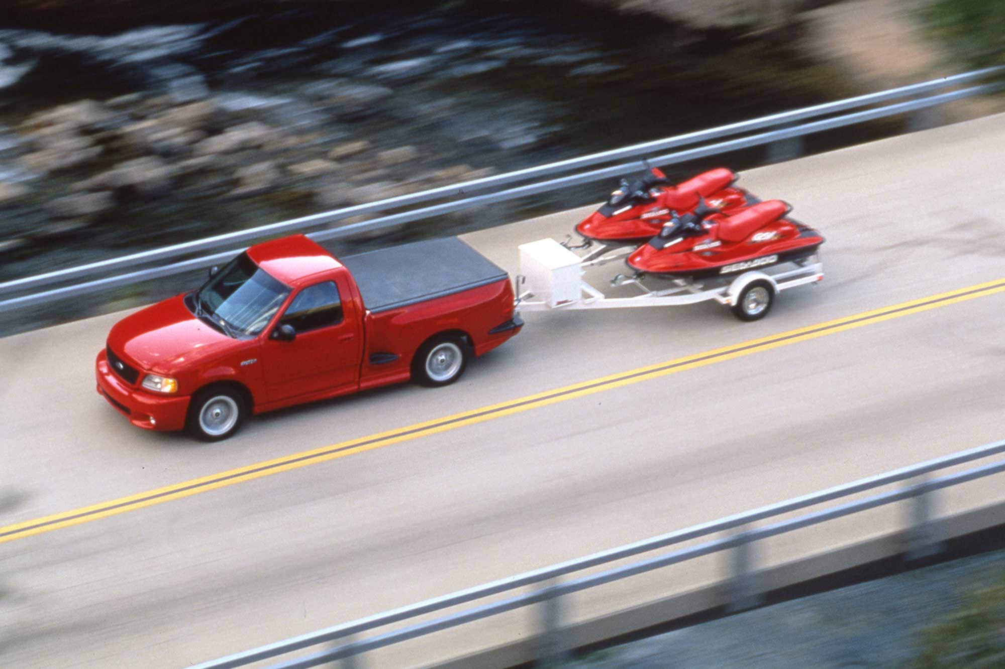 Red 1999 Ford F-150 Lightning towing a pair of personal watercraft on a two-lane bridge.