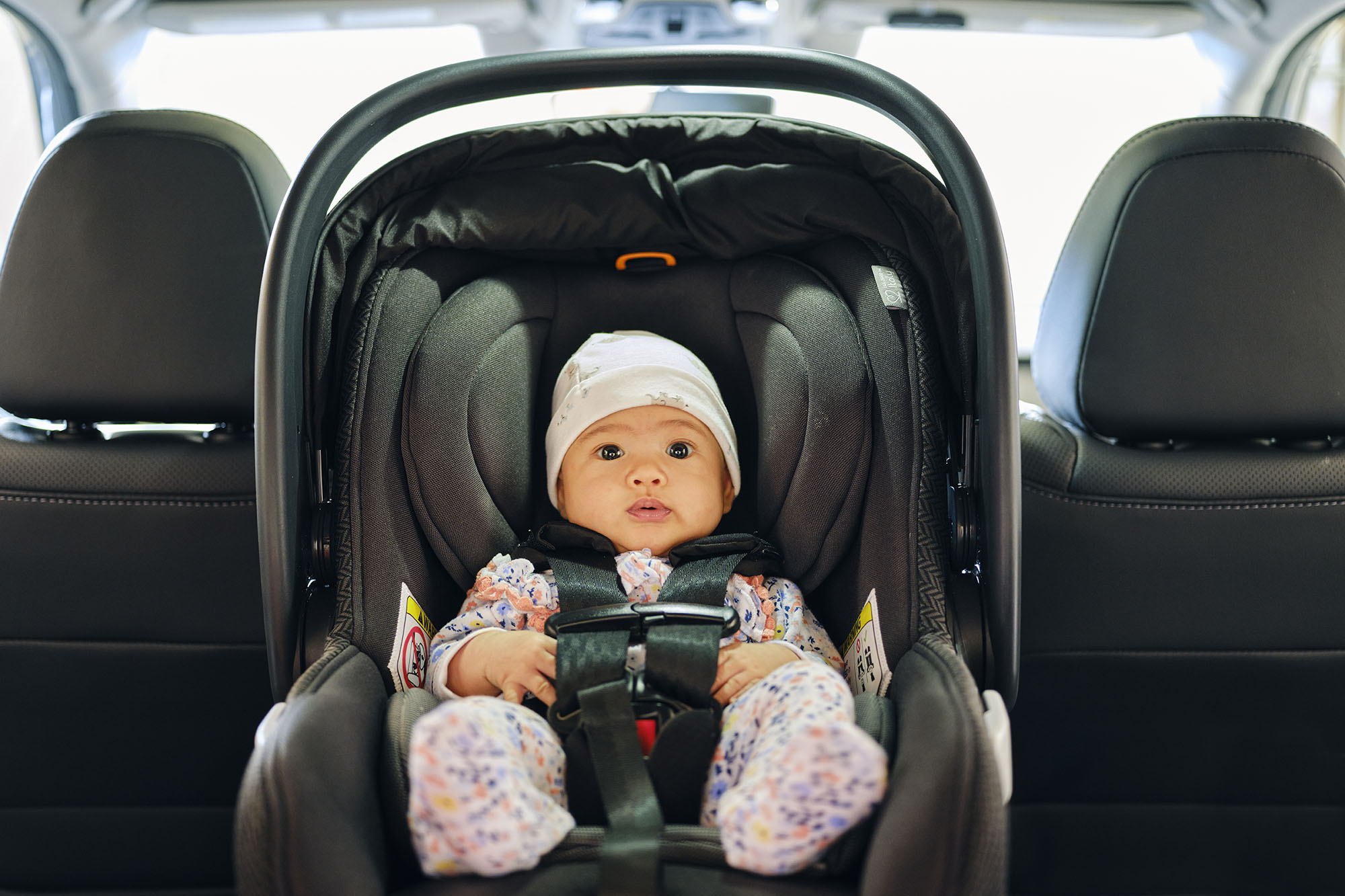 Baby in white hat sits in a rear-facing car seat in the back seat of a vehicle.