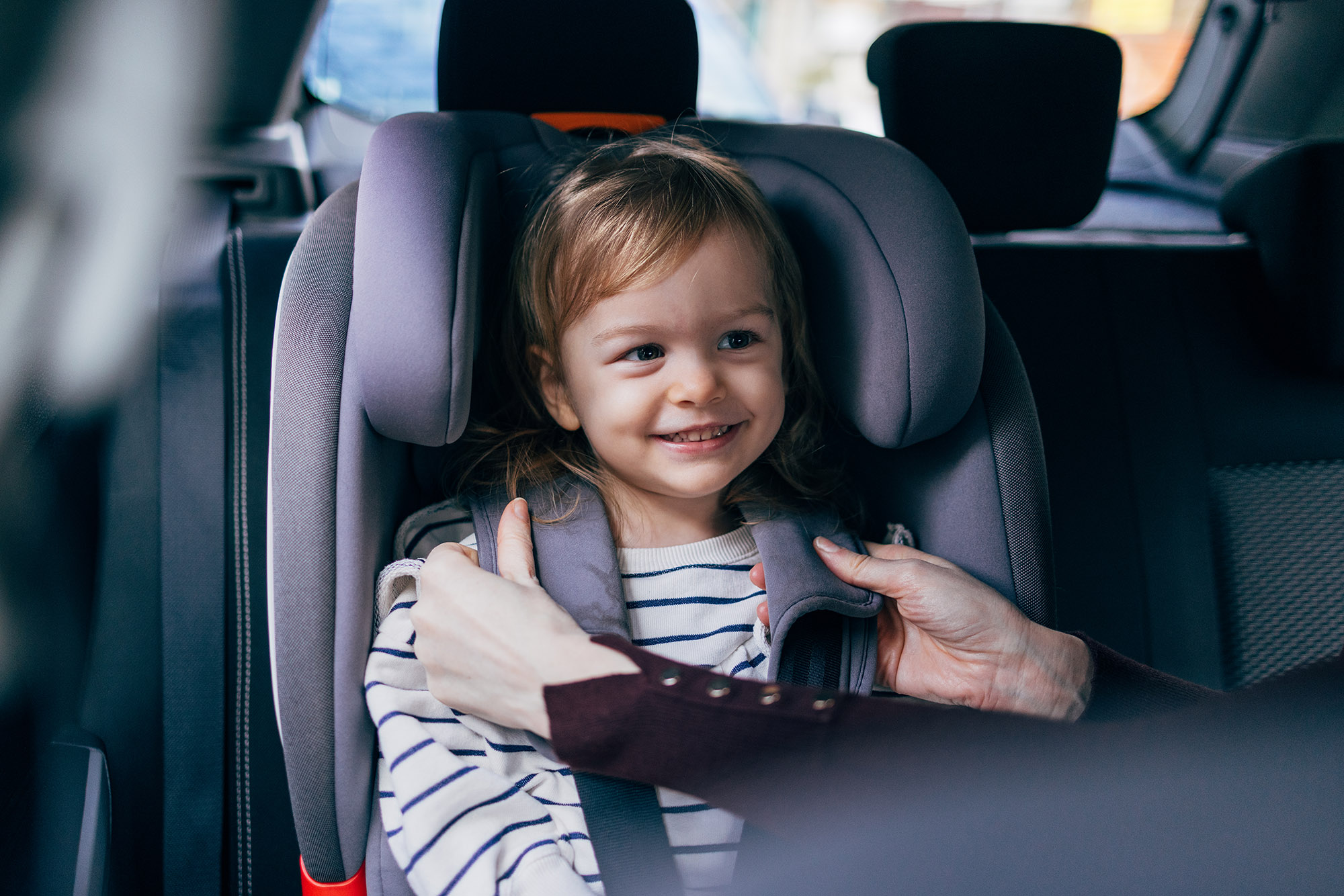 Person buckles smiling child into forward-facing car seat in the back of a vehicle.