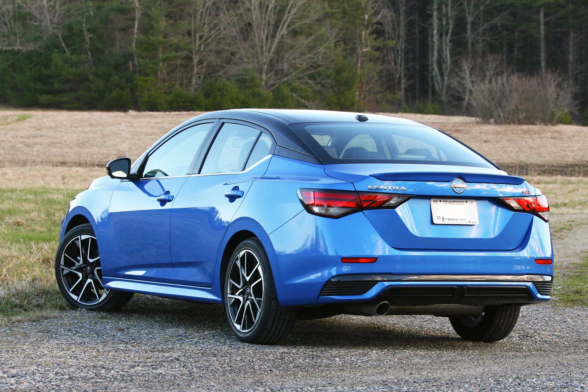 Rear-quarter view of a blue 2024 Nissan Sentra SR parked on gravel with a field and trees in the background.