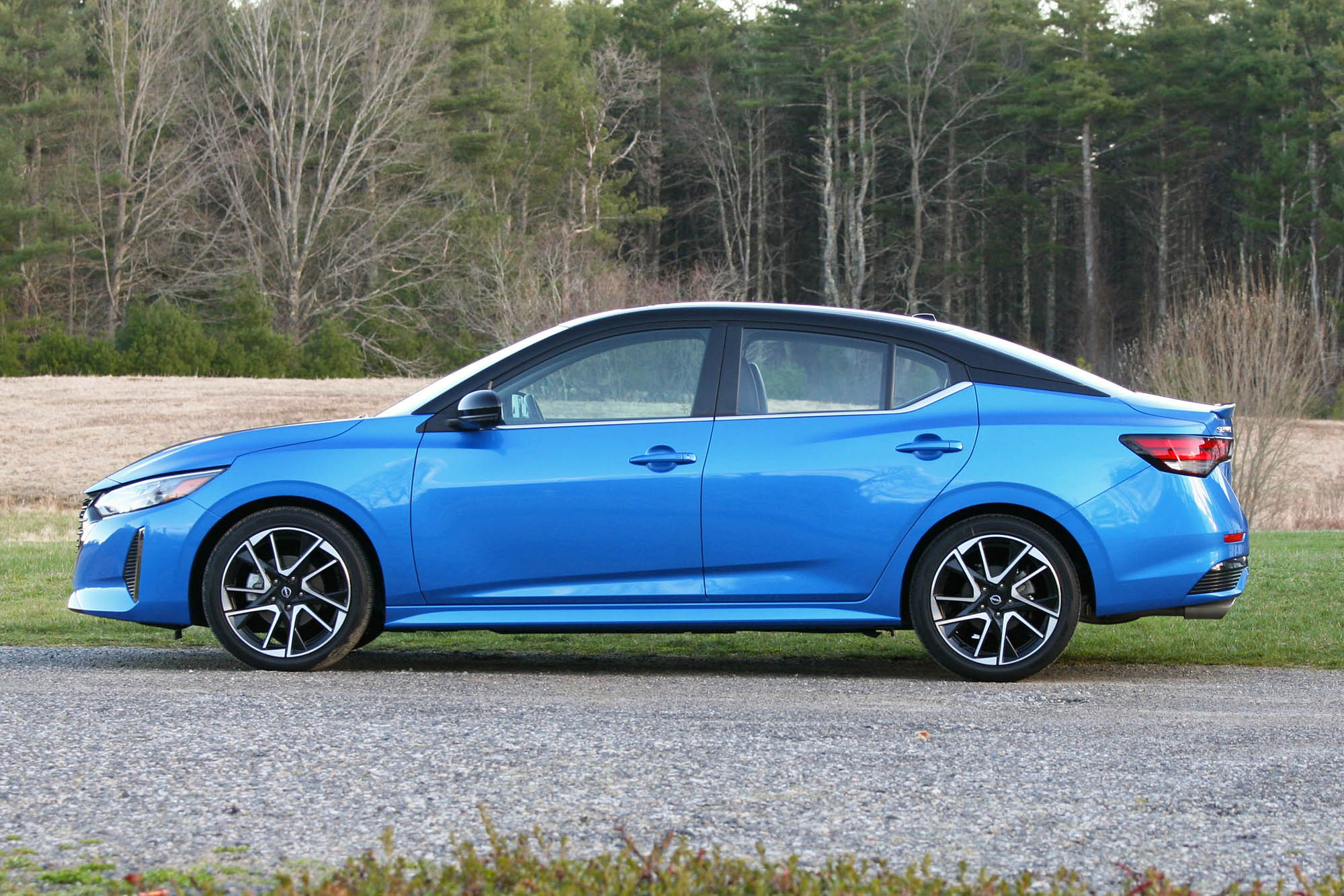 Side view of a blue 2024 Nissan Sentra SR parked on gravel with a field and trees in the background.