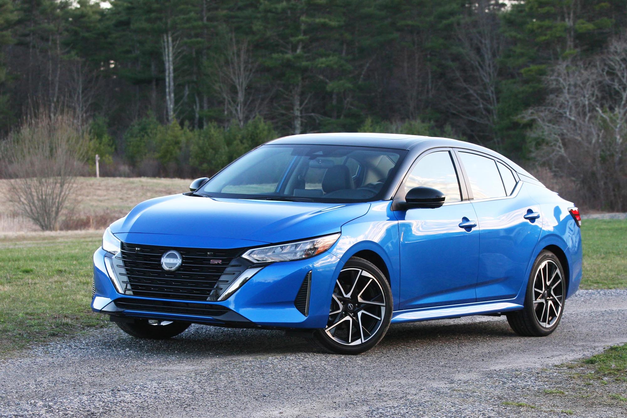 InsFront-quarter view of a blue 2024 Nissan Sentra SR parked on gravel with a field and trees in the background.