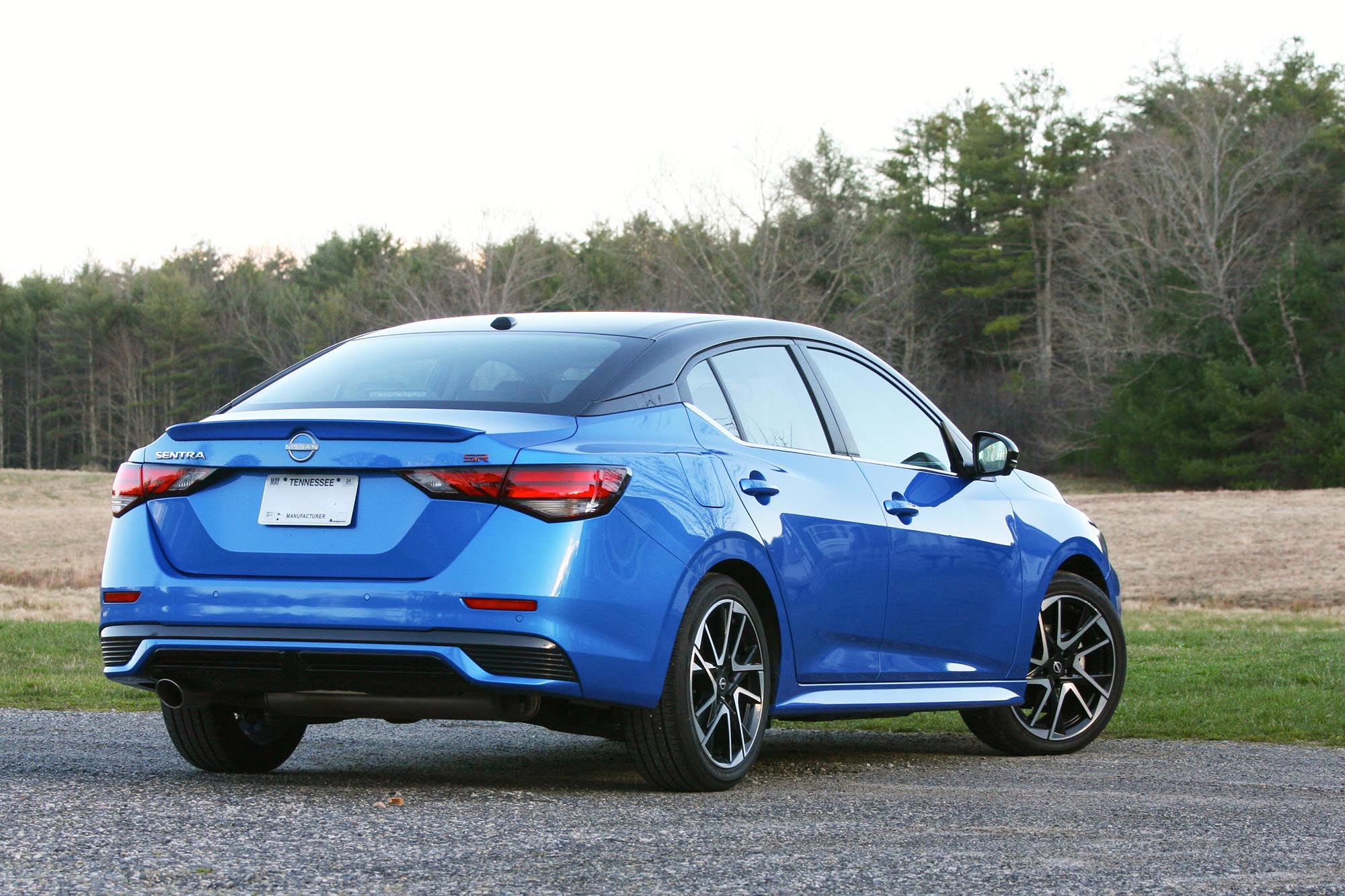 Rear-quarter view of a blue 2024 Nissan Sentra SR parked on gravel with a field and trees in the background.