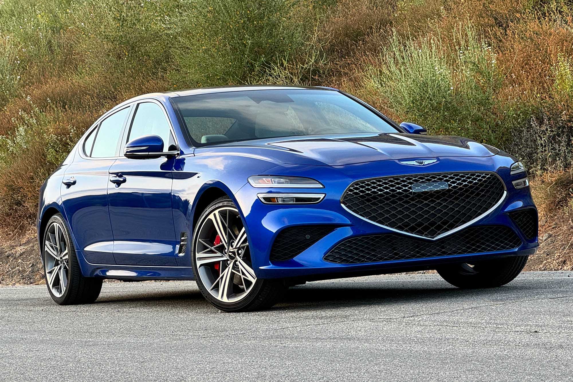 Front-quarter view of a Kawah Blue 2024 Genesis G70 2.5T Sport Prestige parked on pavement with a brush-covered hill behind it.