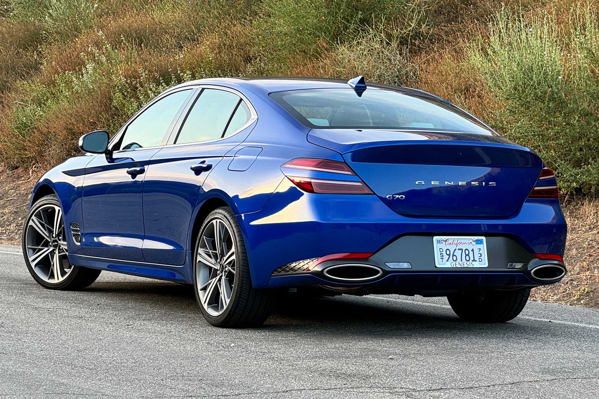 Rear-quarter view of a Kawah Blue 2024 Genesis G70 2.5T Sport Prestige parked on pavement with a brush-covered hill behind it.
