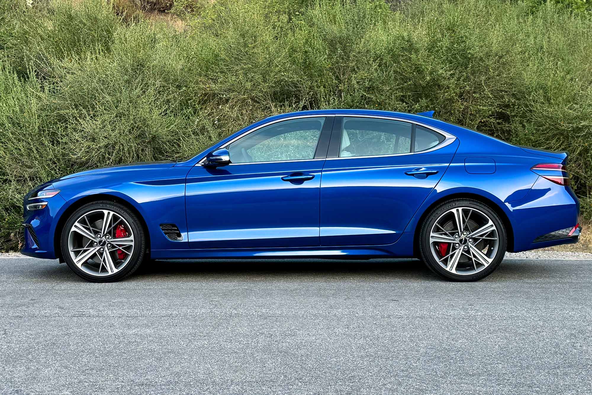 Side view of a blue 2024 Genesis G70 2.5T Sport Prestige parked on pavement with a brush-covered hill behind it