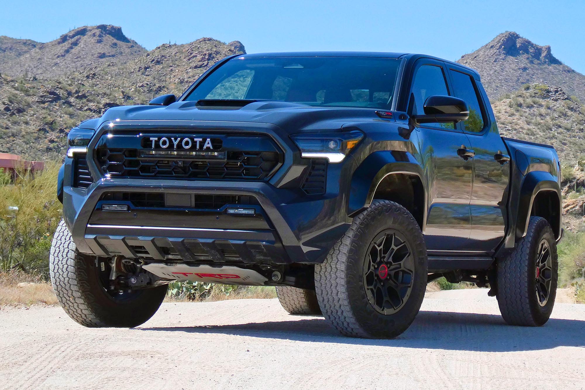 Front-quarter view of a dark gray 2024 Toyota Tacoma TRD Pro parked in the desert with mountains in the background.