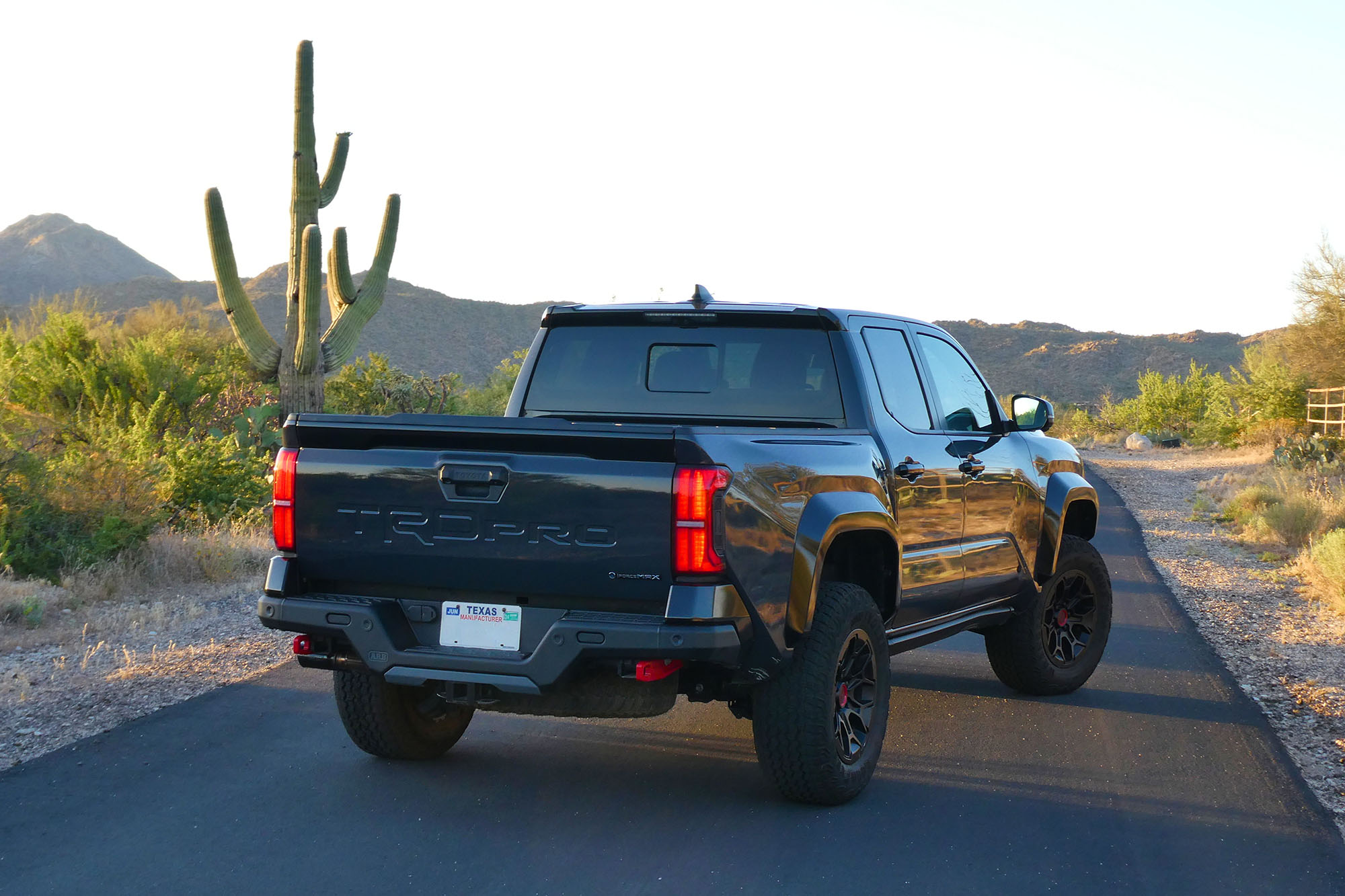 Rear-quarter view of a dark gray 2024 Toyota Tacoma TRD Pro parked on a desert road with a cactus and mountains in the background.