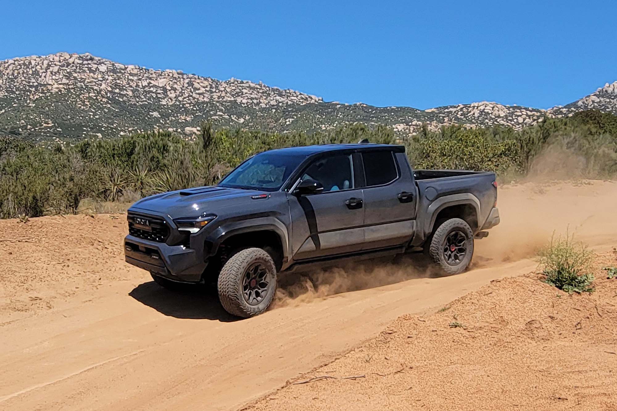 Front-quarter view of a dark gray 2024 Toyota Tacoma TRD Pro driving on a desert trail with rock-covered mountains in the background.