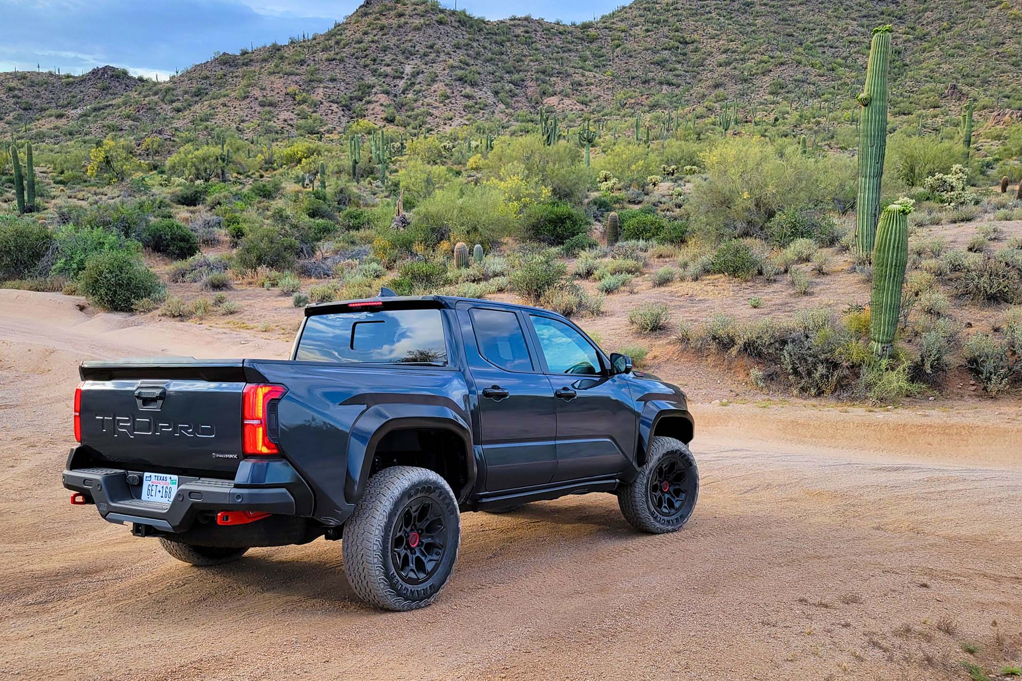 Rear-quarter view of a dark gray 2024 Toyota Tacoma TRD Pro parked in the desert with cacti and mountains in the background.