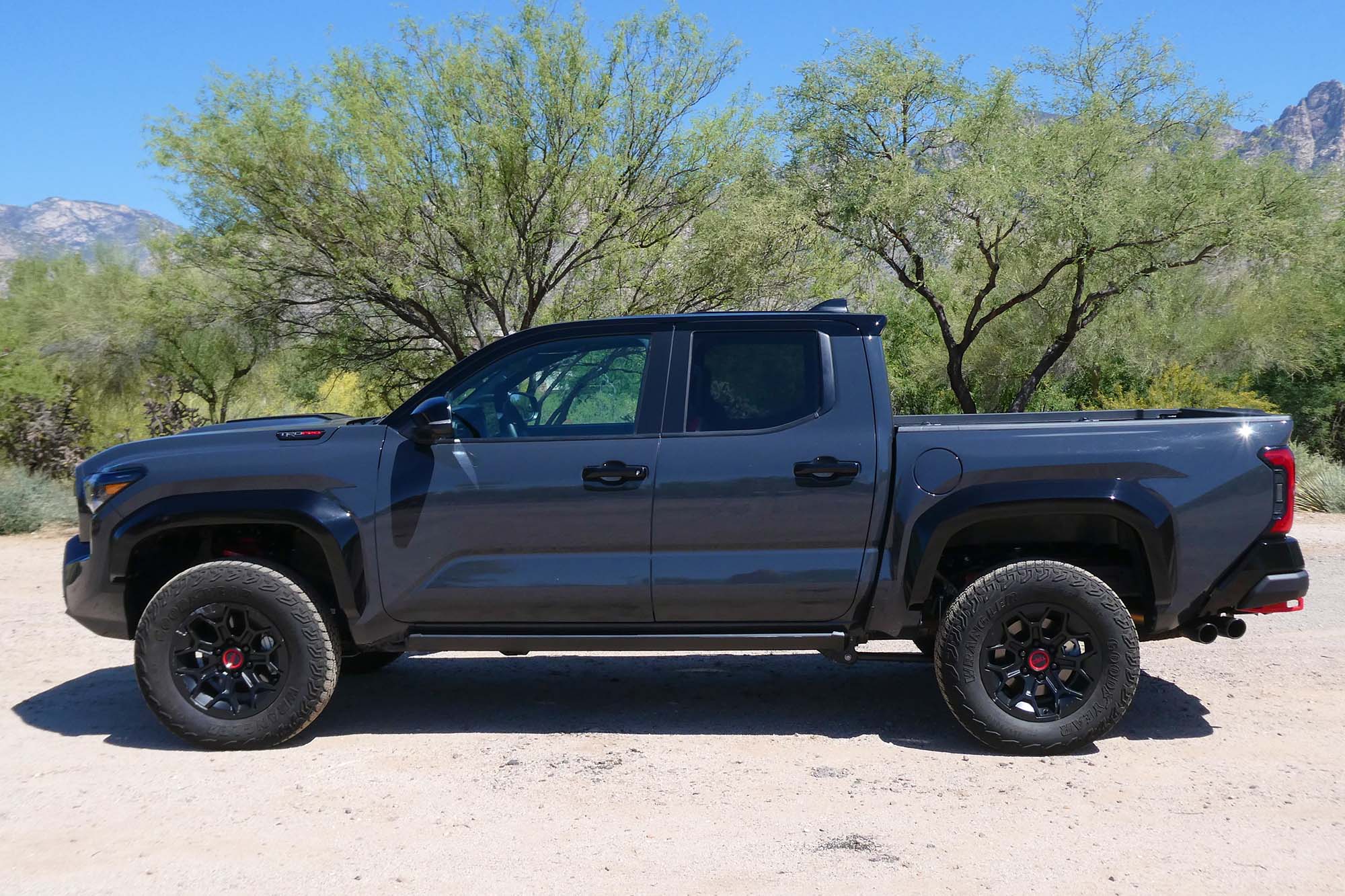 Side view of a dark gray 2024 Toyota Tacoma TRD Pro parked in the desert with trees and mountains in the background.