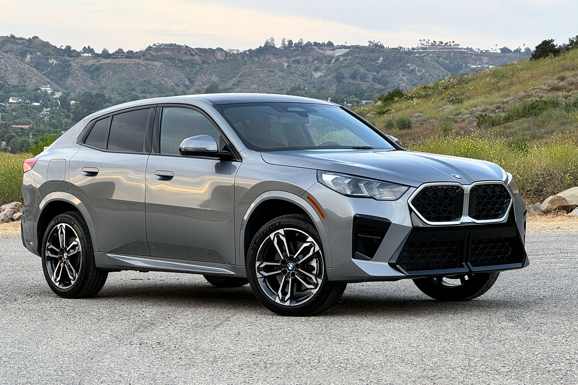 Gray 2024 BMW X2 parked on pavement in front of hills and a grassy slope.