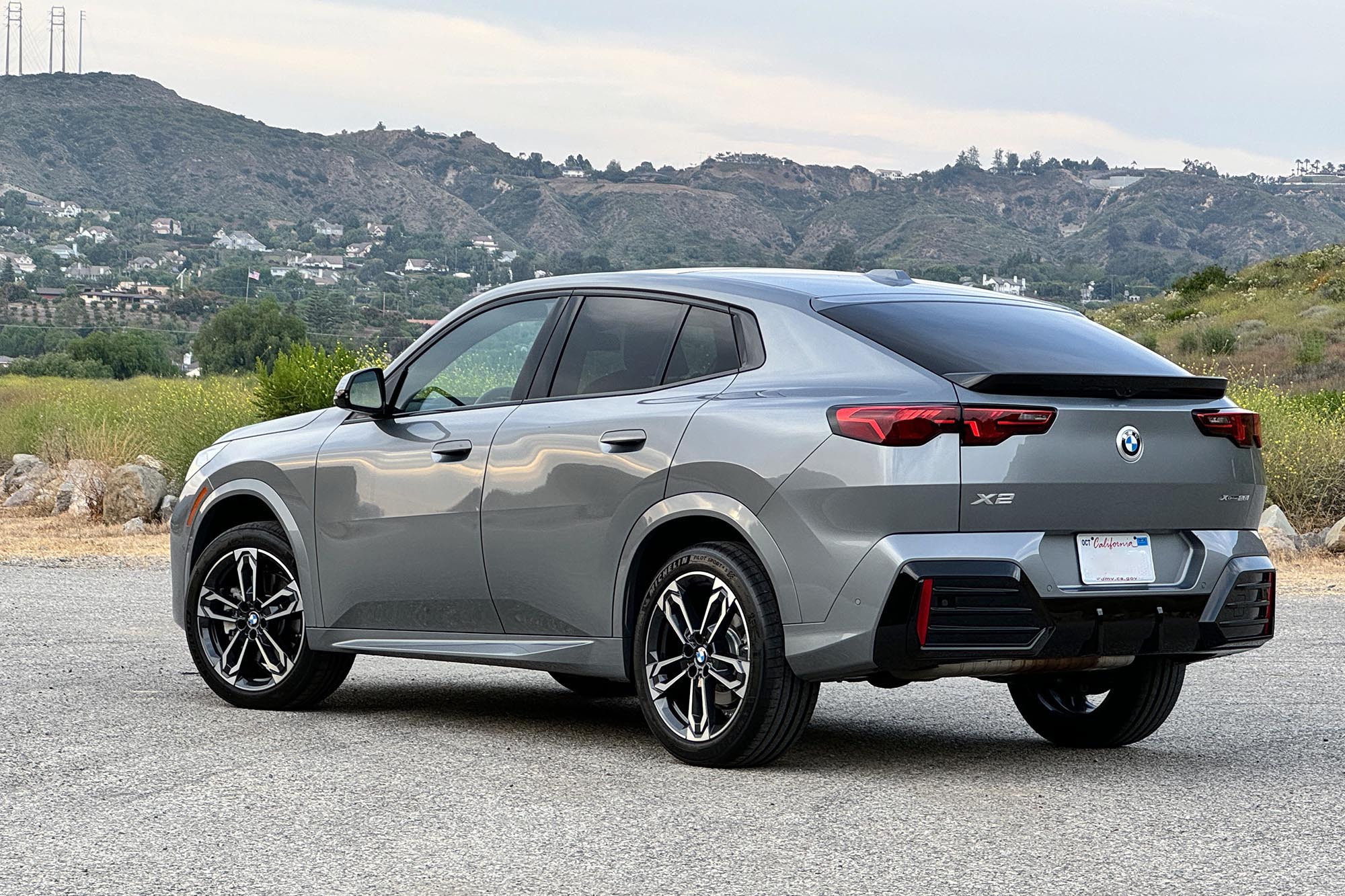 Gray 2024 BMW X2 parked on pavement in front of hills and a grassy slope.