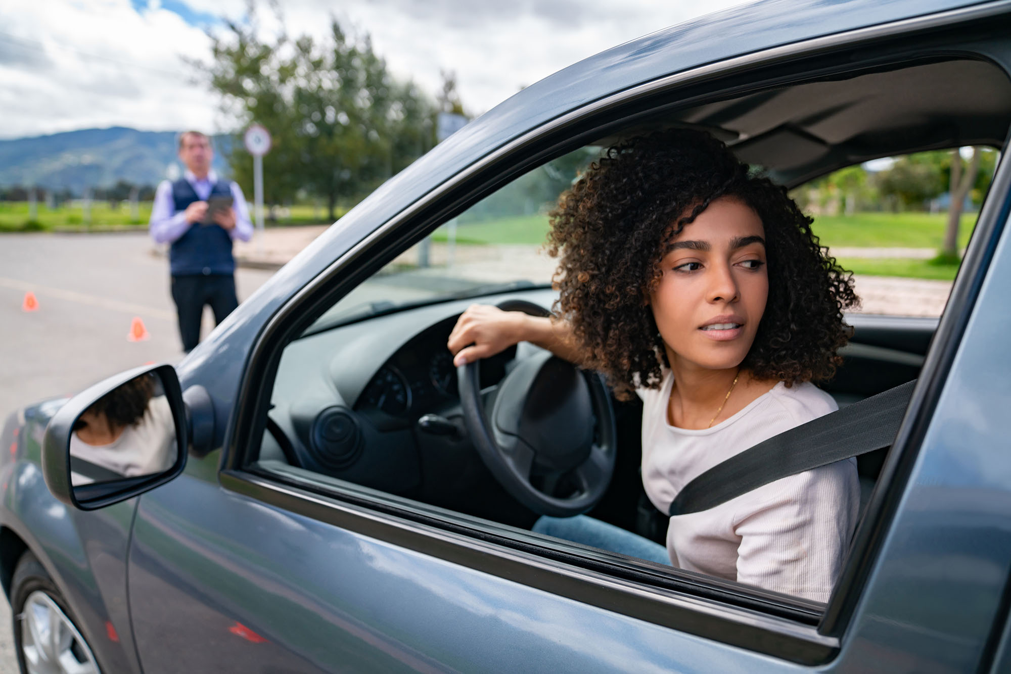 Young driver looks out the car window to parallel park with a driving instructor in the background.