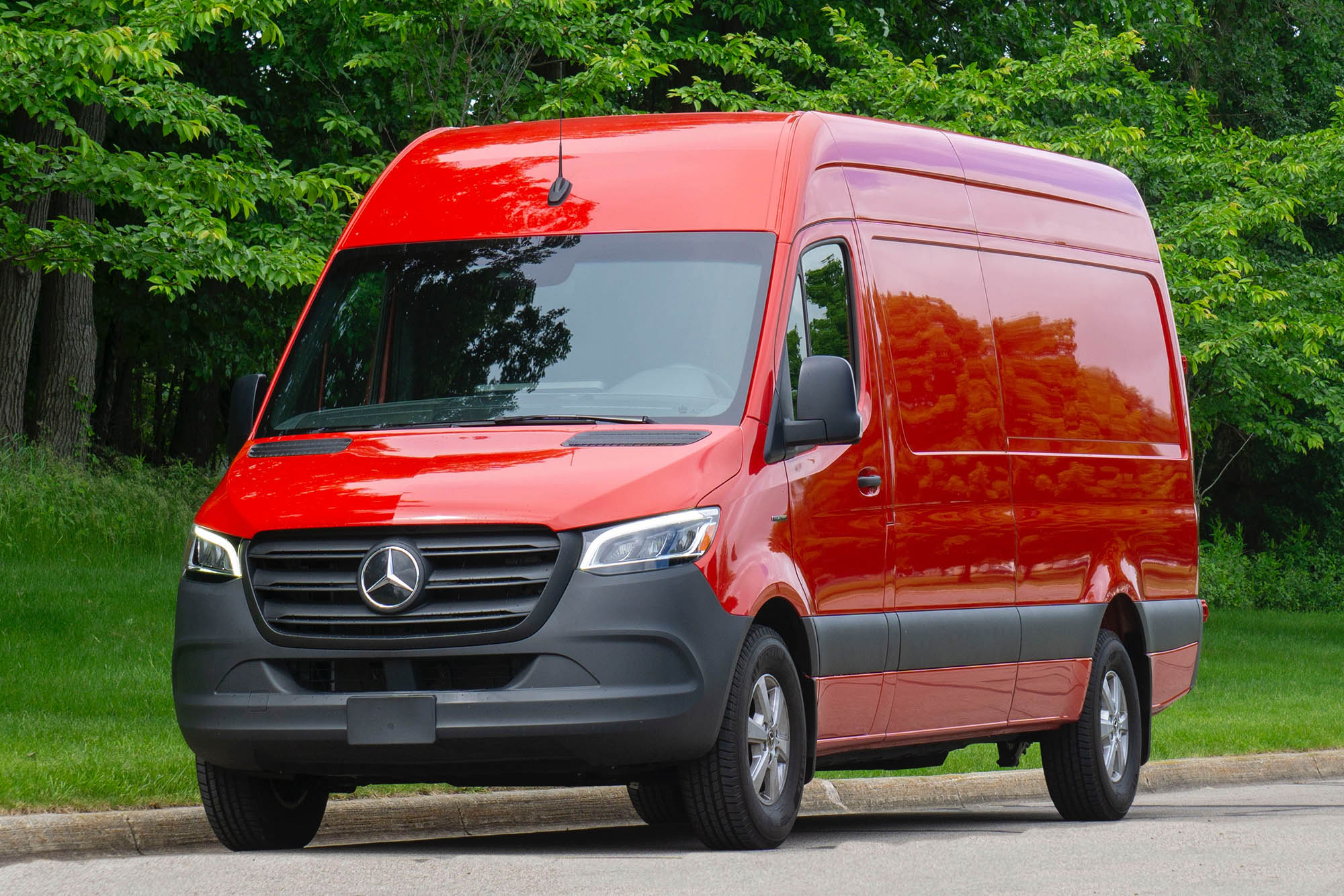 A front-quarter view of a red 2024 Mercedes-Benz eSprinter parked on a street with trees in the background