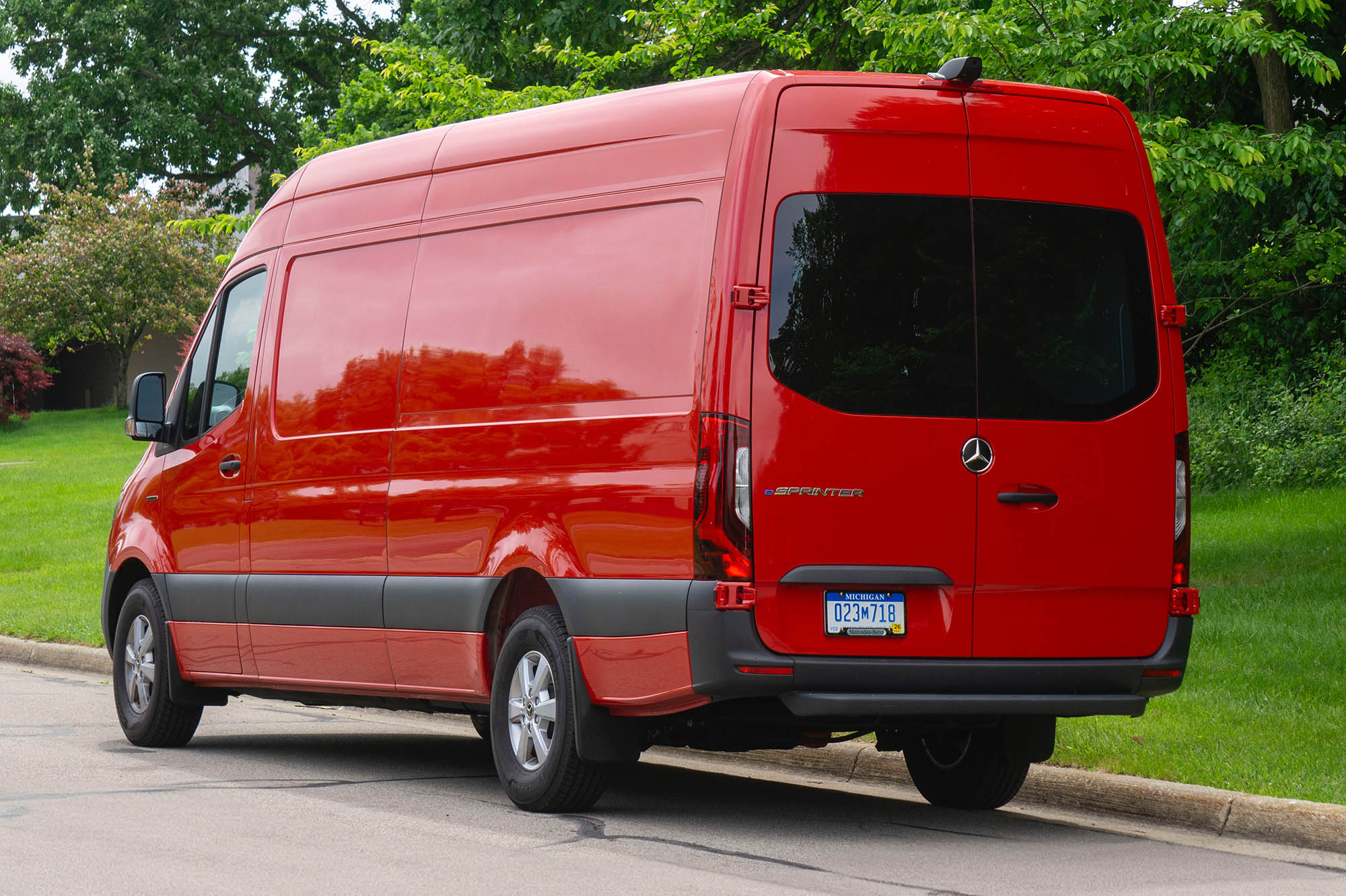 A rear-quarter view of a red 2024 Mercedes-Benz eSprinter parked on a street with trees in the background