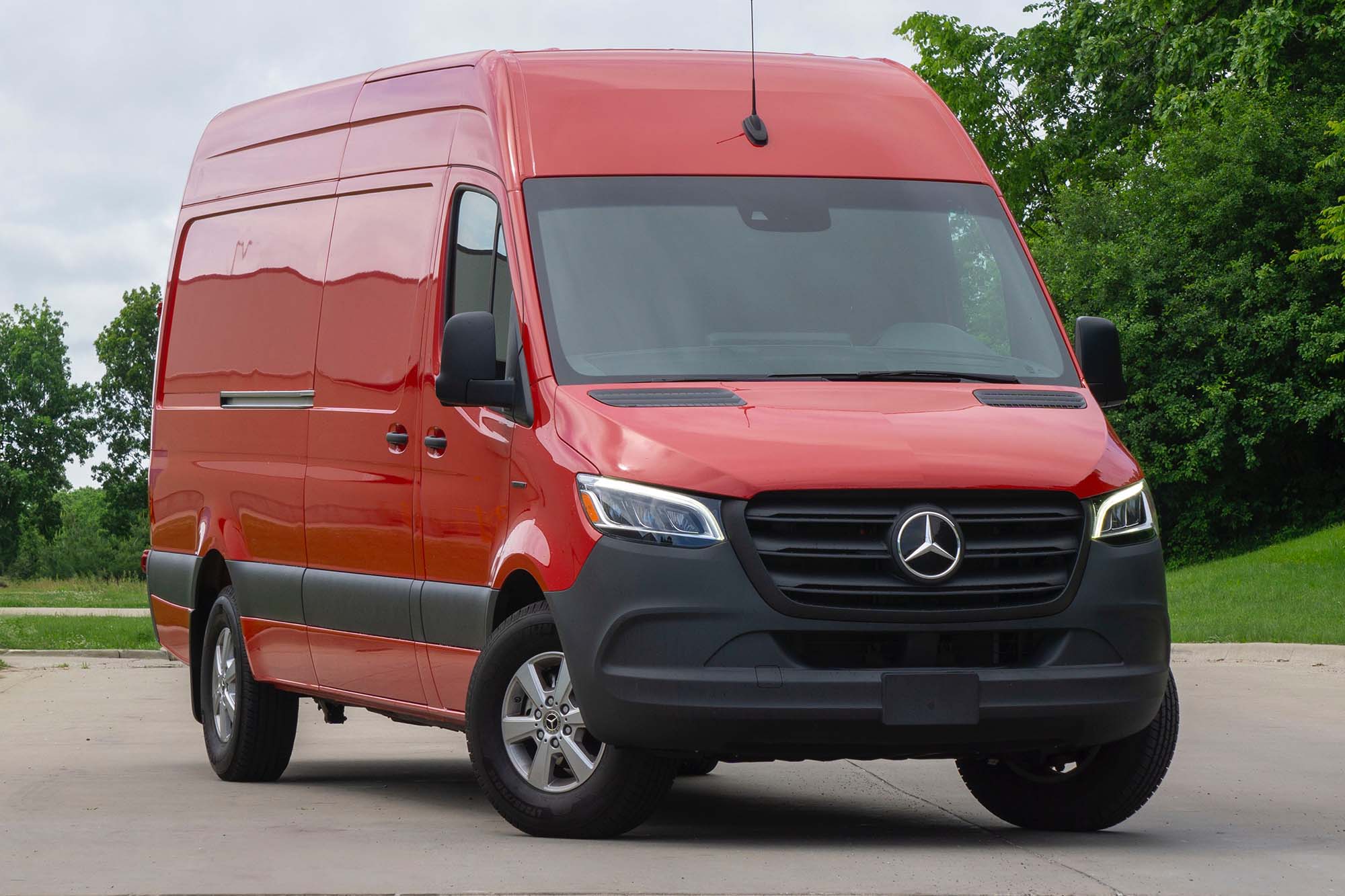 A front-quarter view of a red 2024 Mercedes-Benz eSprinter parked on a street with trees in the background