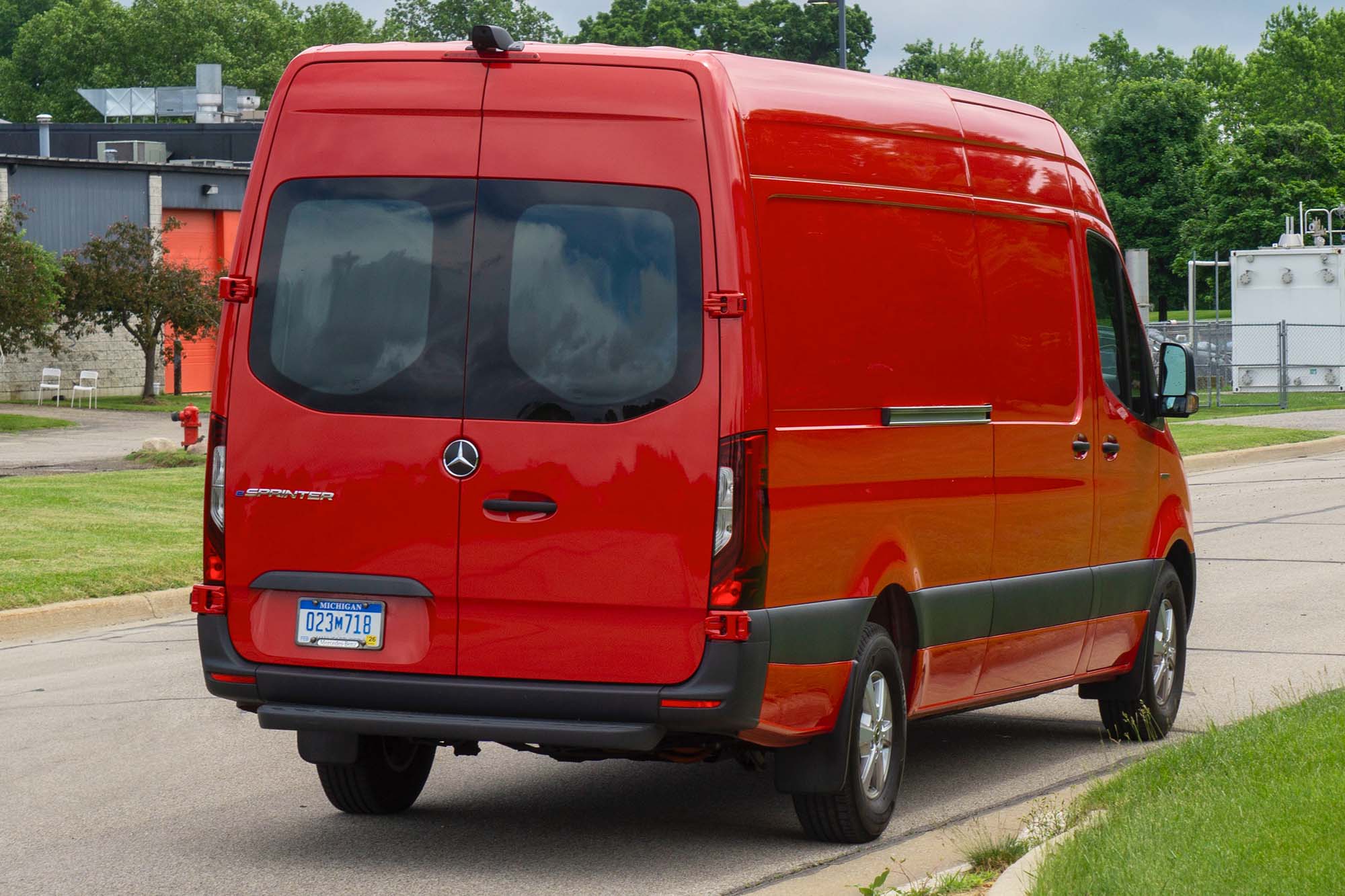 A rear view of a red 2024 Mercedes-Benz eSprinter parked on a street with industrial facilities in the background