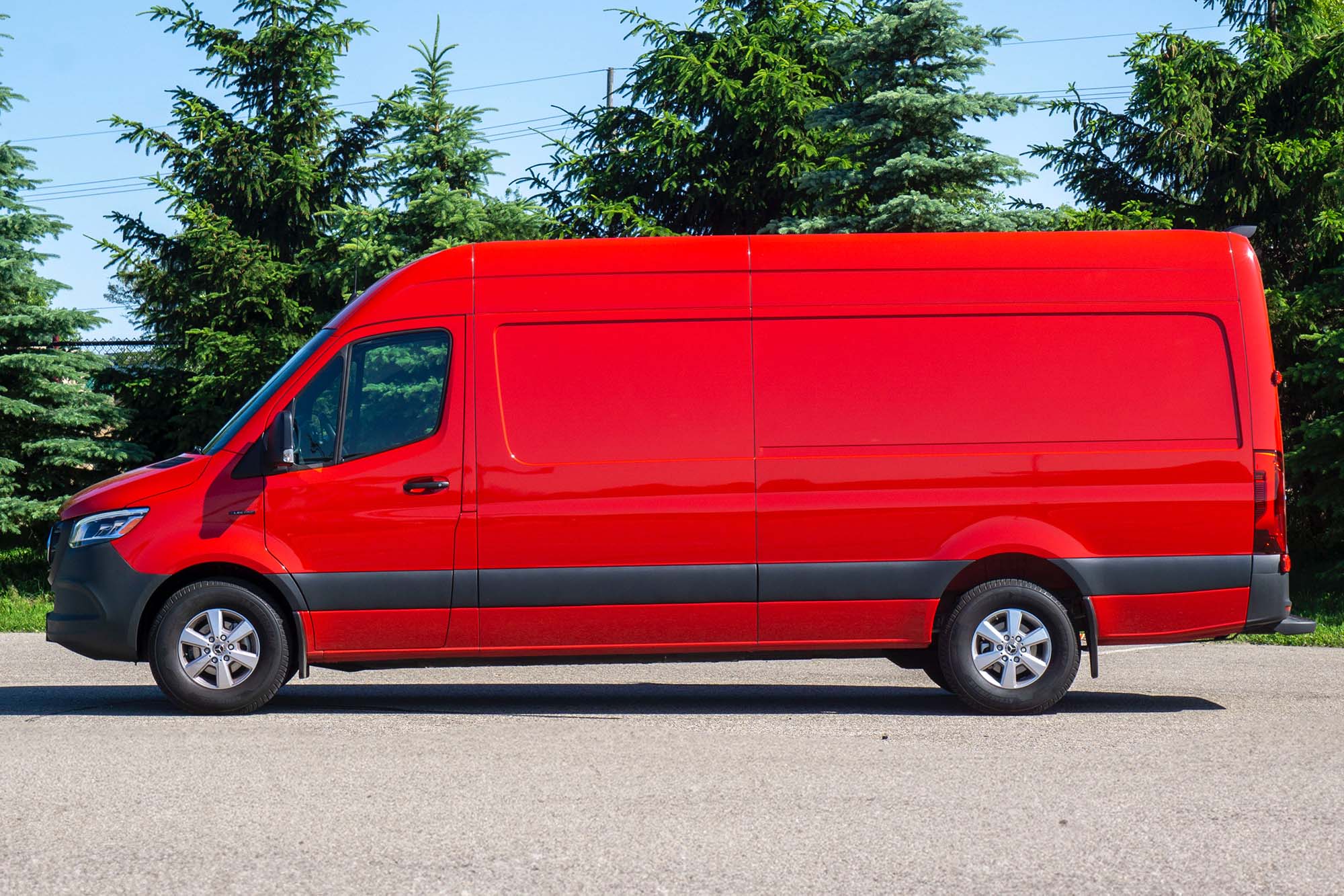 A side view of a red 2024 Mercedes-Benz eSprinter parked on the pavement with trees in the background