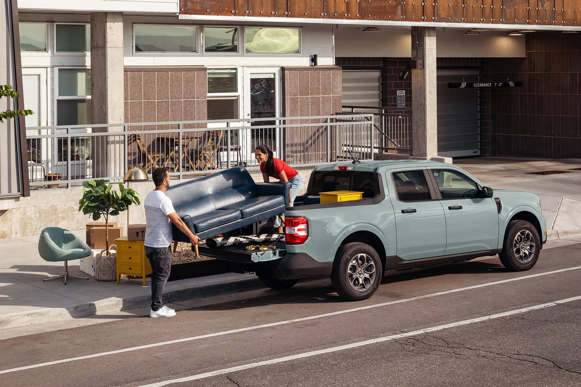 Two people load a couch into the rear of a light blue Ford Maverick