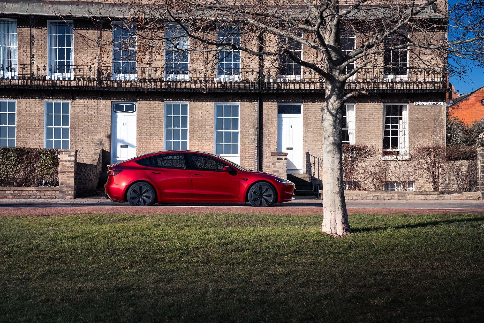 A red Tesla Model 3 with aero wheels parked in front of a brick building