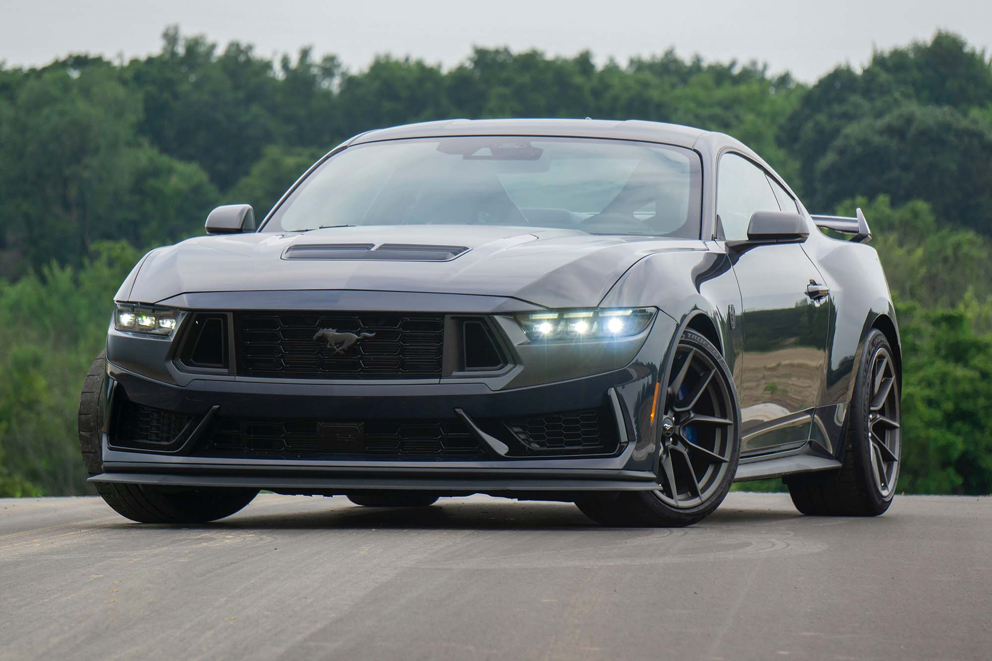 Front-quarter view of a Blue Ember Metallic 2024 Ford Mustang Dark Horse parked on blacktop with trees in the background.