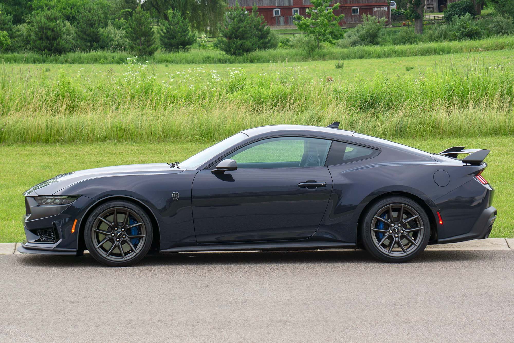 Side view of a Blue Ember Metallic 2024 Ford Mustang Dark Horse parked on a road with a field and an old red barn in the background.