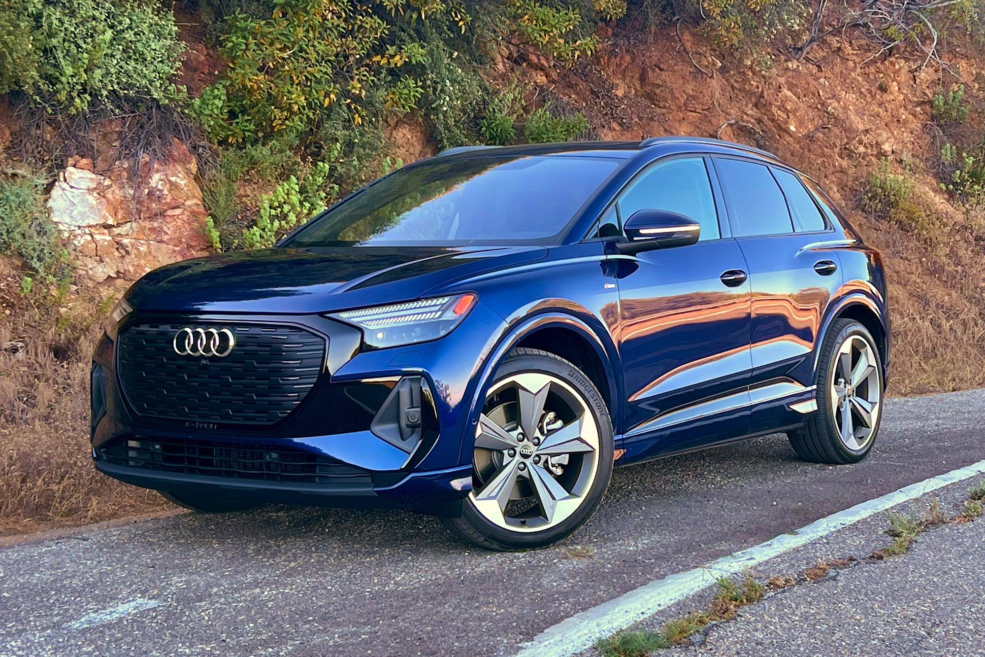 Front-quarter view of a blue 2024 Audi Q4 55 e-tron parked on the side of a road with a hillside behind it.