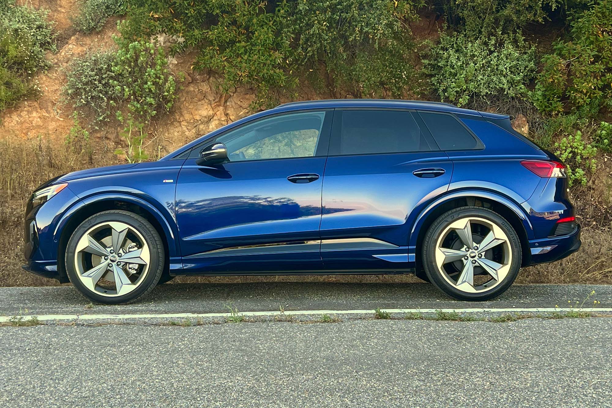 Side view of a blue 2024 Audi Q4 55 e-tron parked on the side of a road with a hillside behind it.