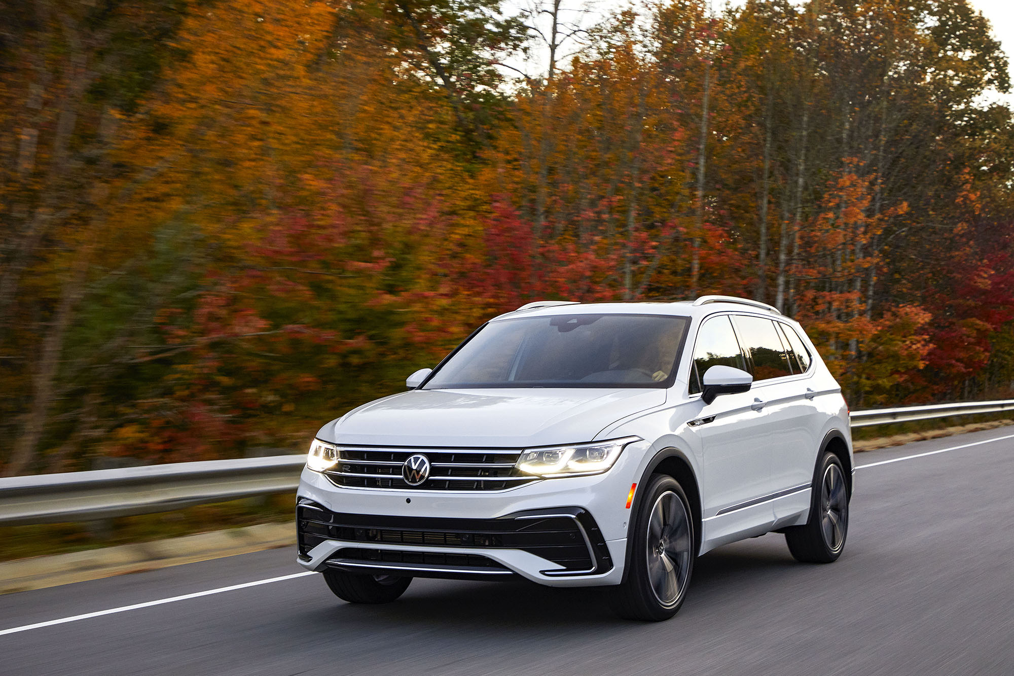 White Volkswagen Tiguan driving on paved road with autumn foliage in the background.