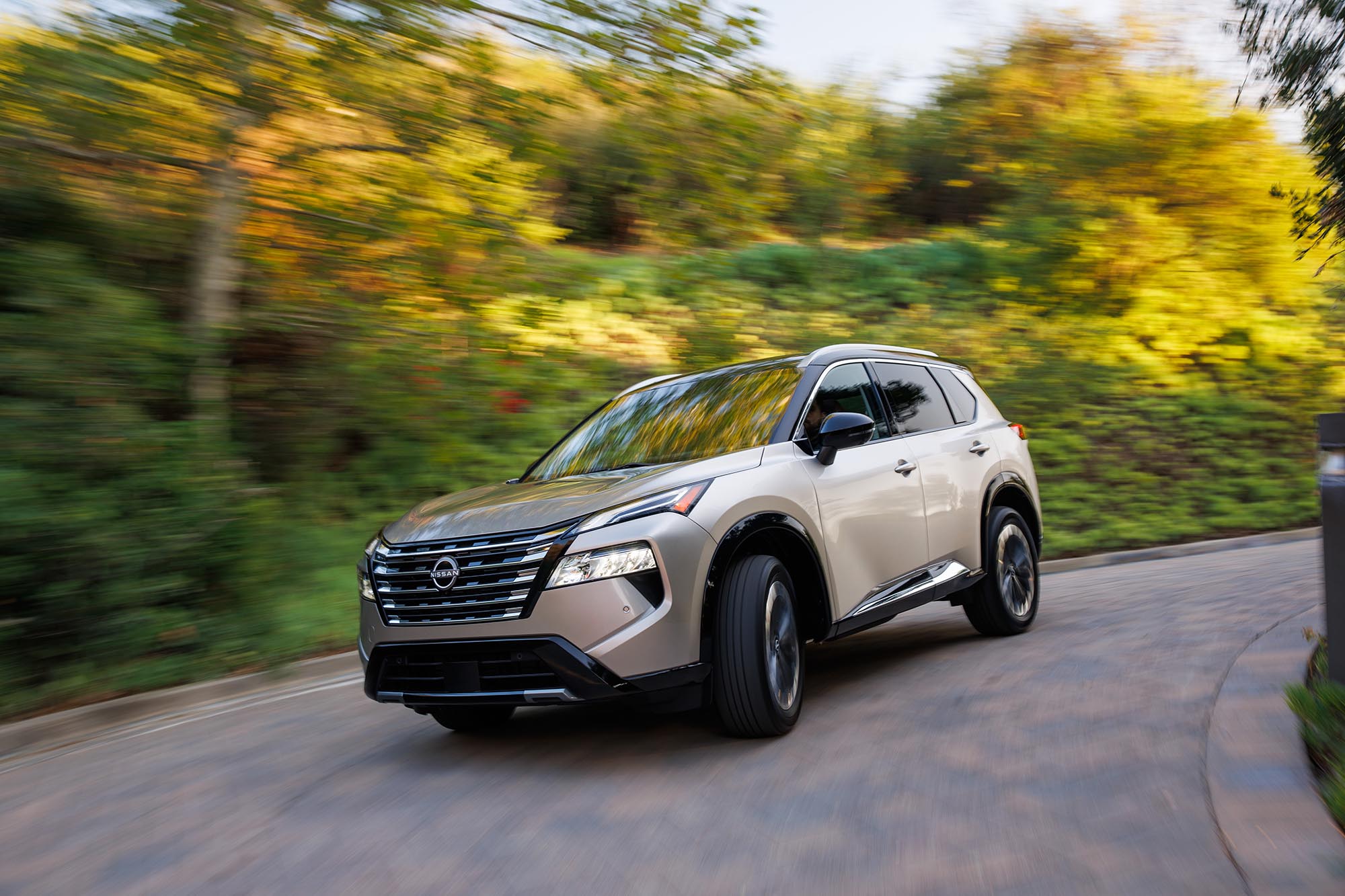 Tan Nissan Rogue driving on paved road with green foliage in the background.